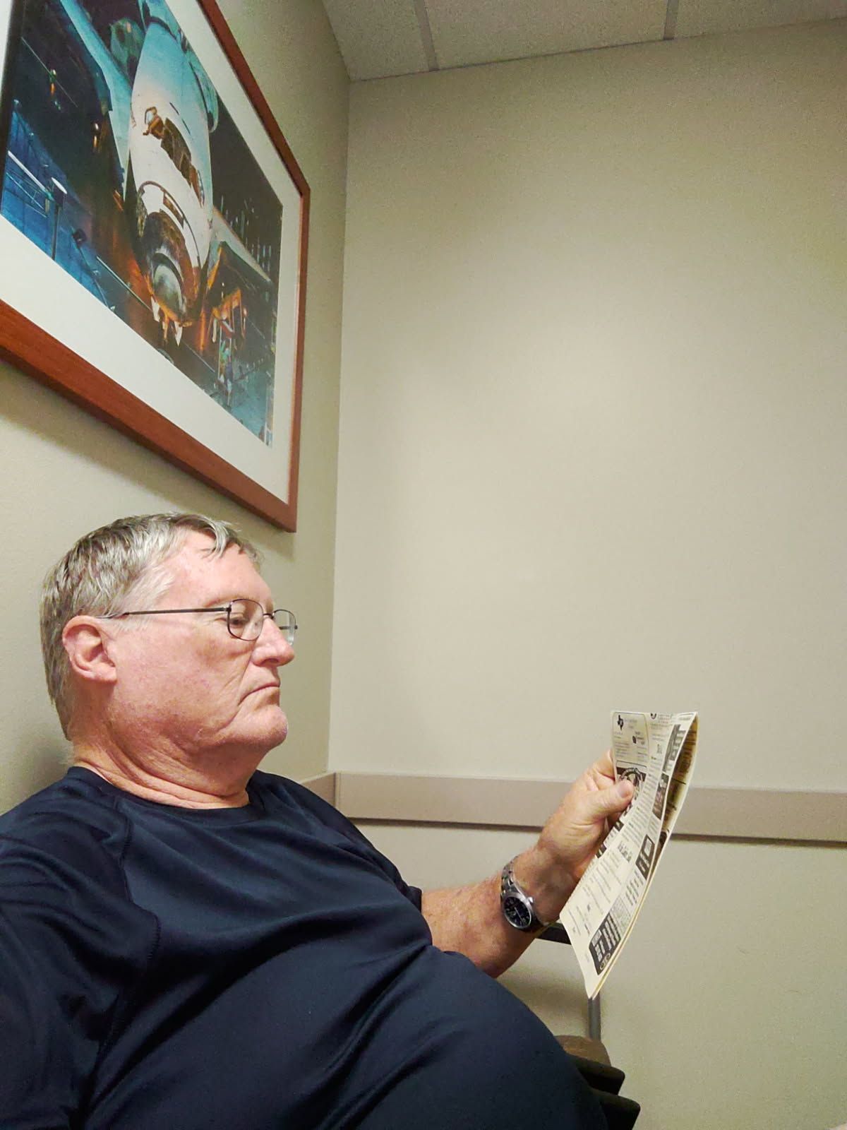 Man in glasses, reading a paper, seated in a waiting area with a framed artwork on the wall.