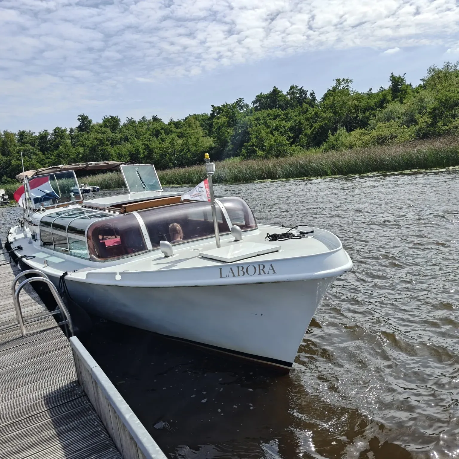 Een lichtgekleurde kajuitboot genaamd TABORA meerde aan bij een houten steiger aan een rivier, vlakbij een met bomen omzoomde oever.