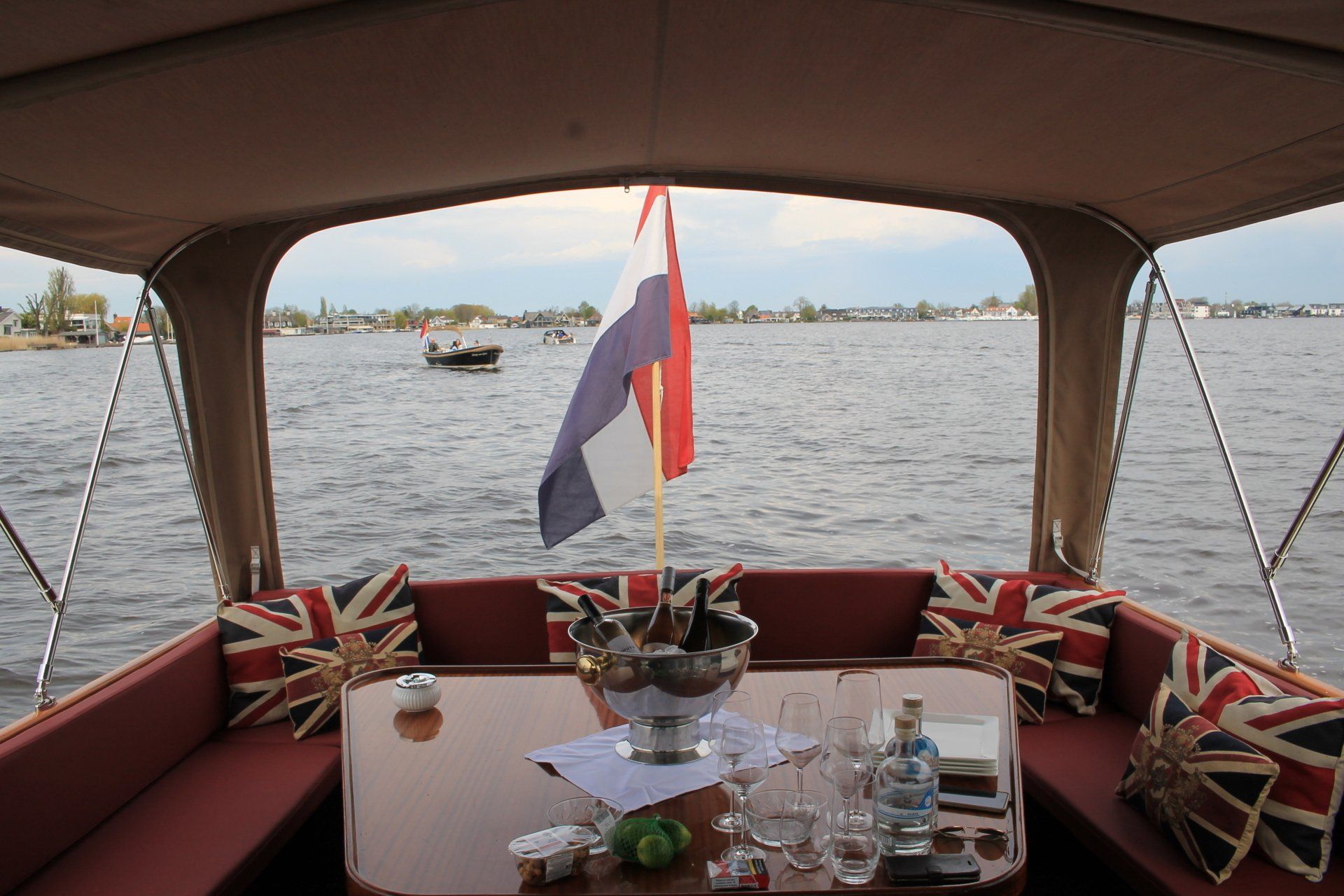 Achteraanzicht vanaf een bootdek onder een luifel, met een tafel met drankjes en kussens met de Britse vlag, met uitzicht op een rivier.