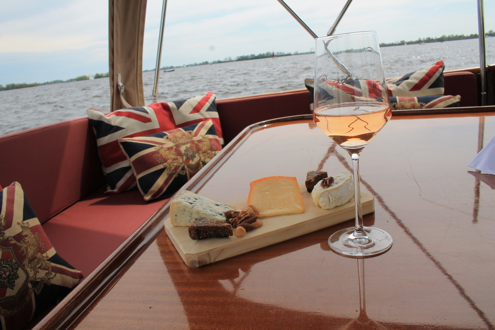 Een glas roséwijn en een kaasplank met diverse kazen op een gepolijste tafel op een boot, met kussens met de Britse vlag op de achtergrond.