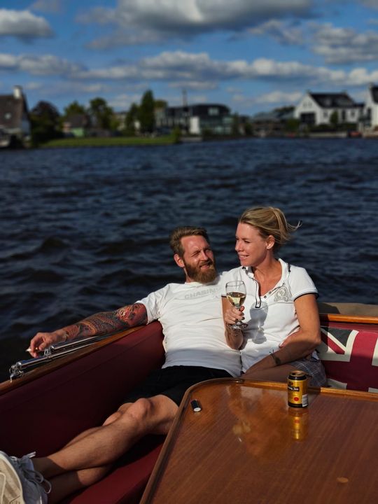 Een stel ontspant met een drankje op een houten boot op een meer onder een zonnige hemel, met huizen op de achtergrond.