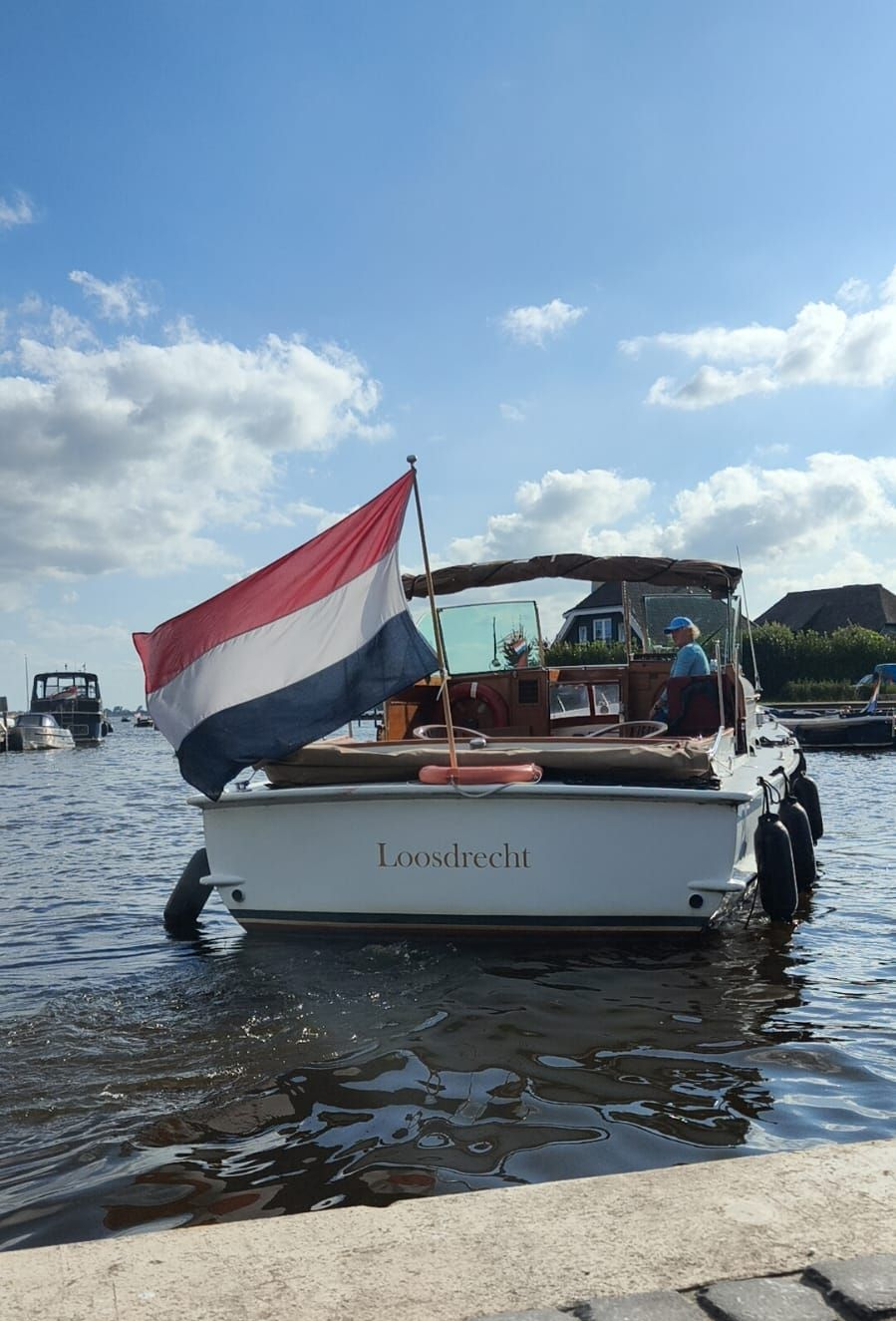 Een witte motorboot met een Nederlandse vlag op de achtersteven ligt aangemeerd aan een stenen kade onder een heldere, zonnige hemel.