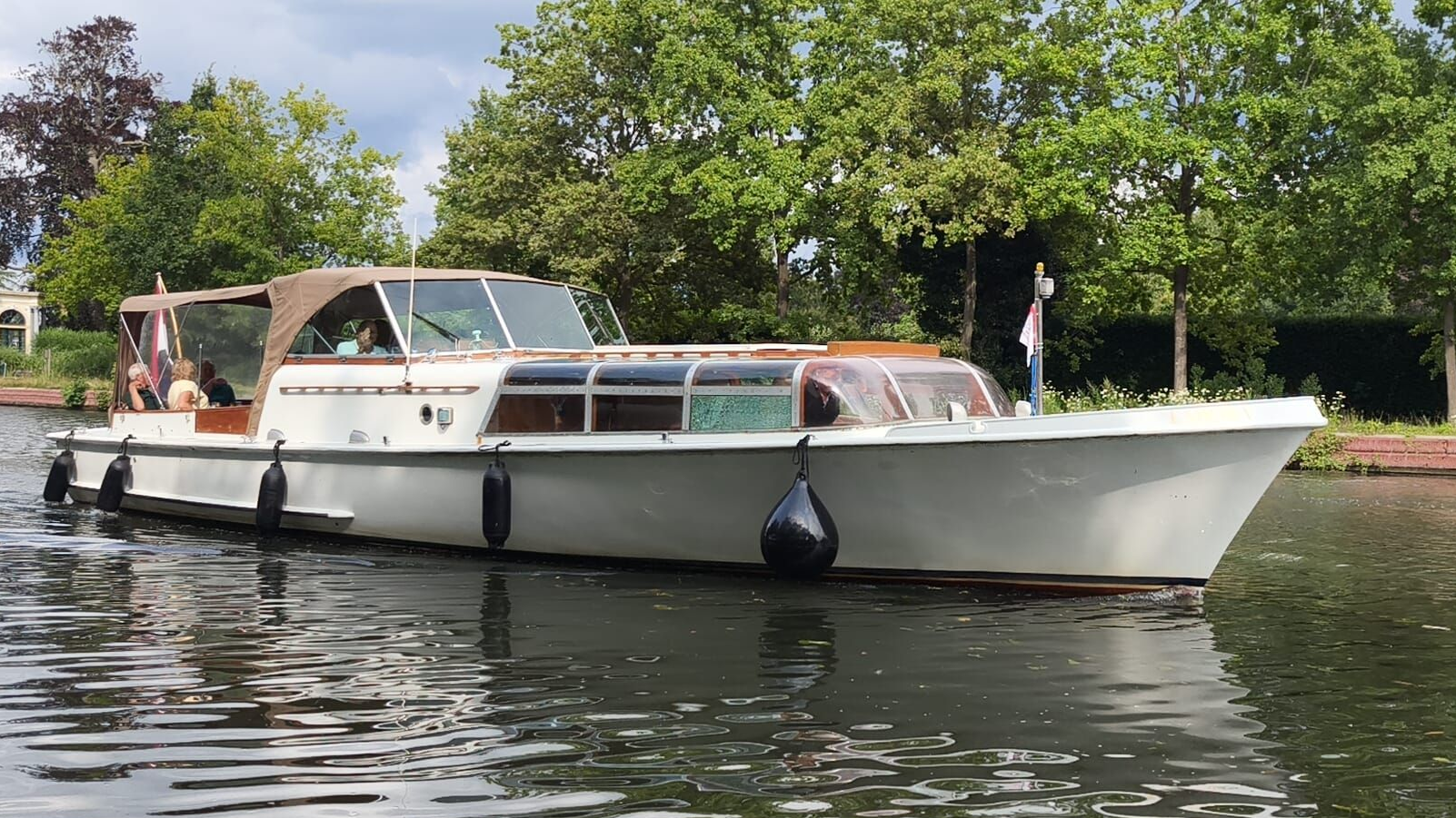 Een witte motorboot met een beige kap vaart over een kalme rivier, omzoomd door groene bomen.