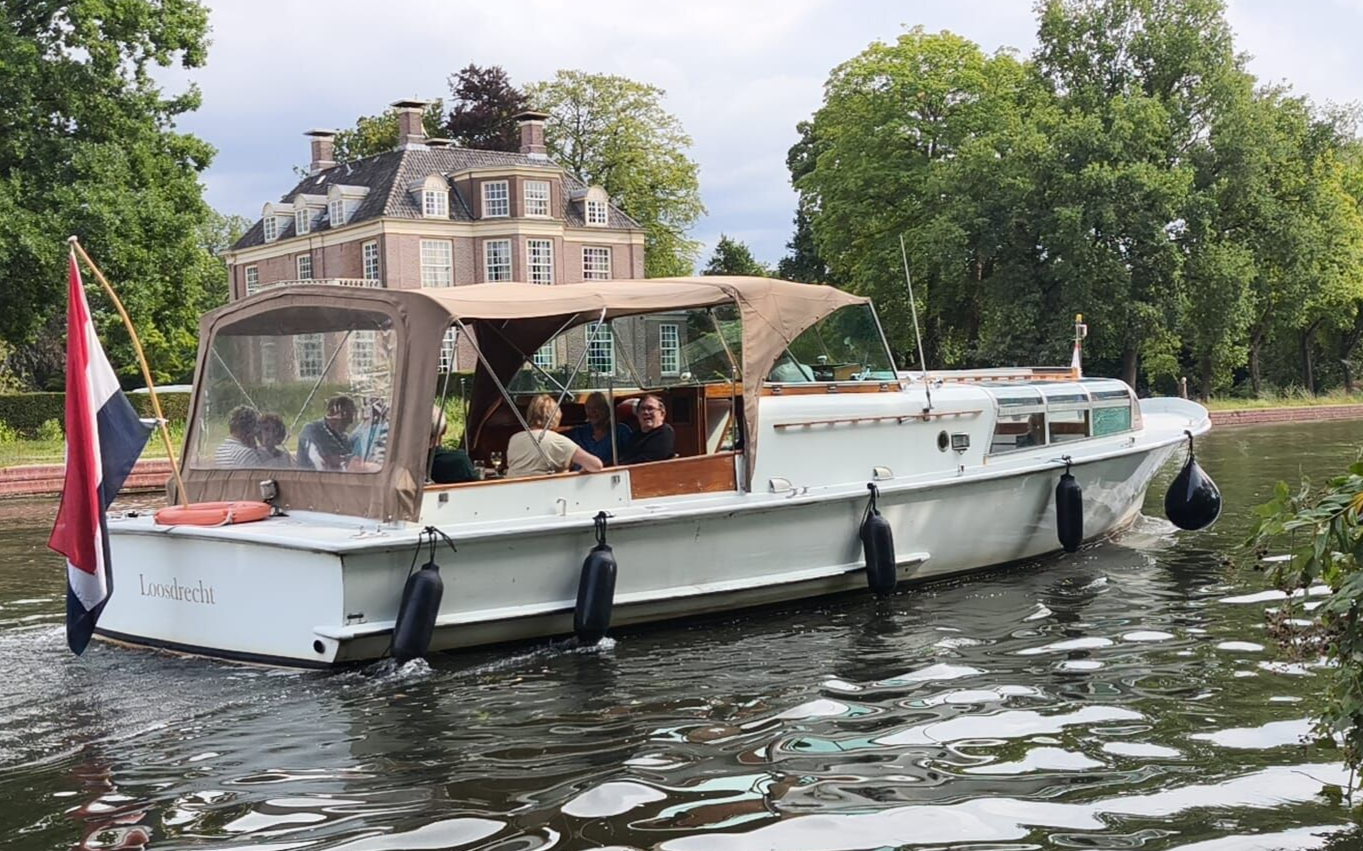 Een witte motorboot met een beige overkapping vervoert passagiers over een rustig kanaal en vaart langs een groot huis aan een groene, lommerrijke oever.