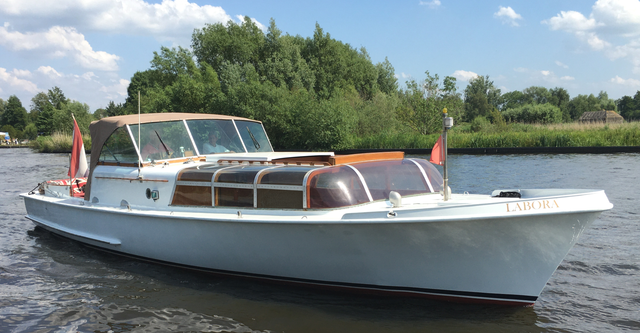 Een witte motorboot met een bruin canvas dak en glazen dekramen vaart over een rivier die omzoomd is door groene bomen.
