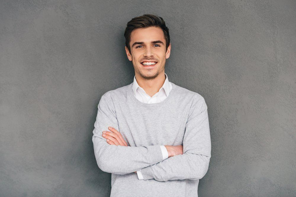 A Man in A Grey Sweater Is Standing with His Arms Crossed and Smiling — Cane to Coast Dentures in Proserpine, QLD