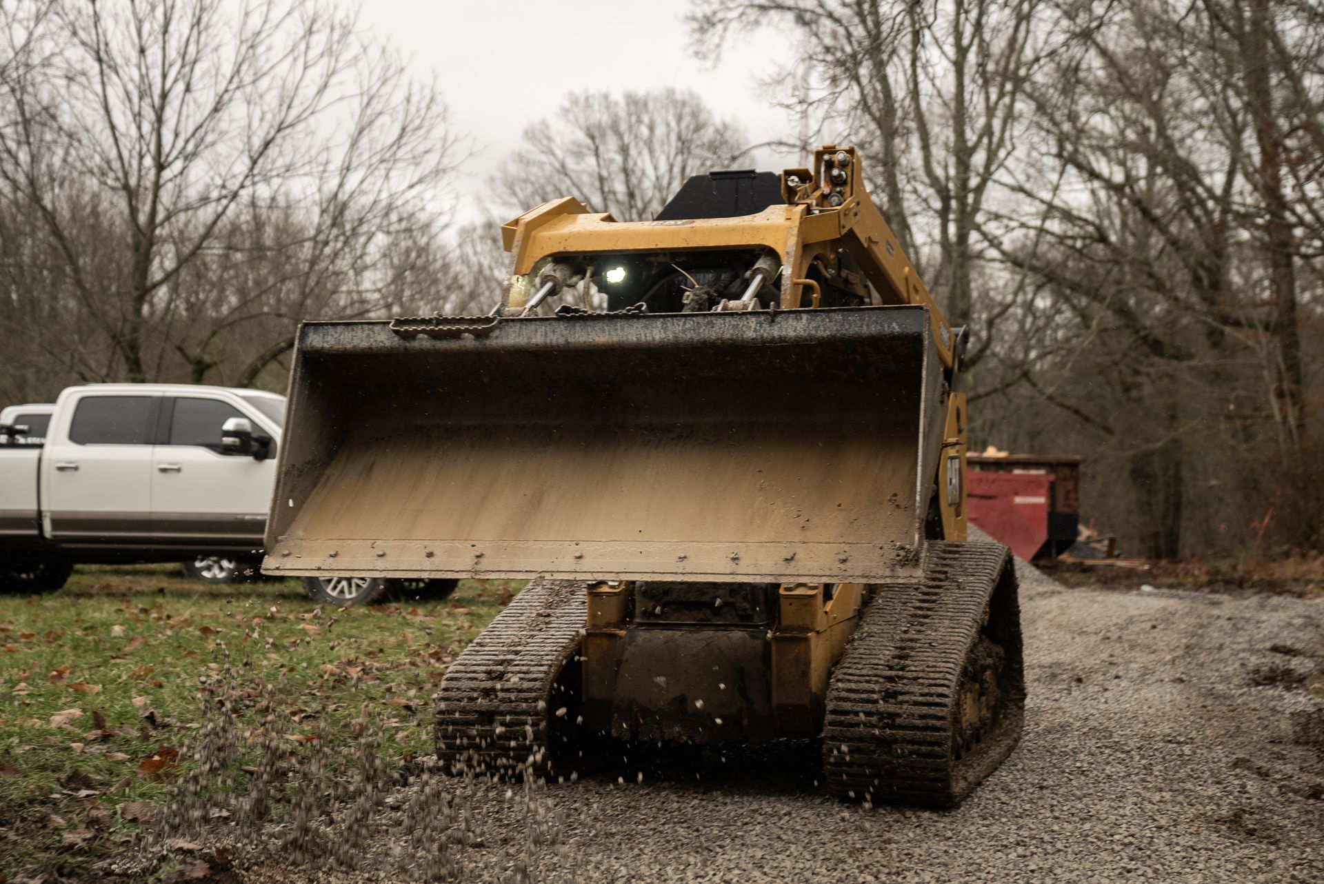 Yellow track loader scooping gravel on a dirt path, white truck in background, overcast day.