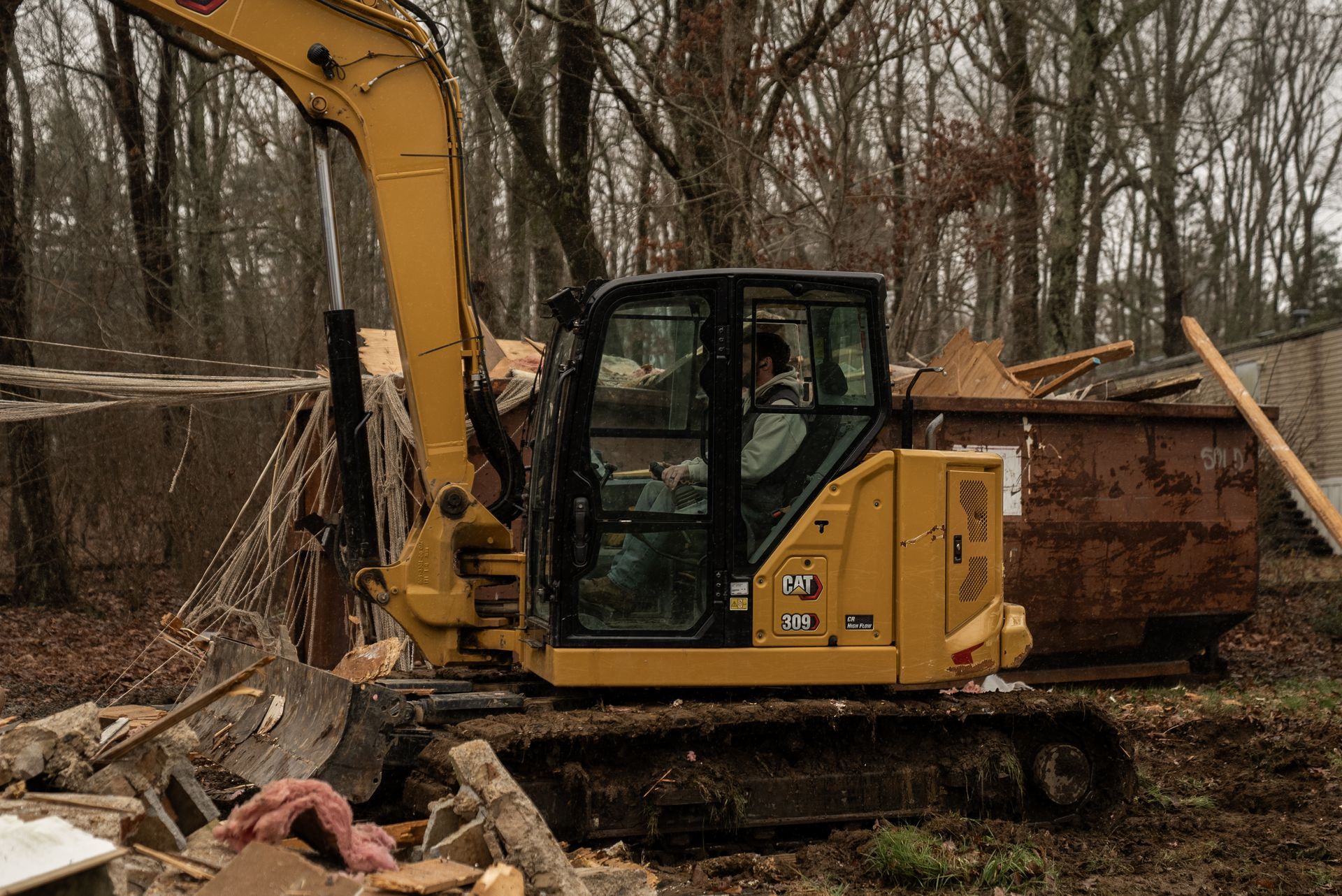 Yellow excavator removing debris near a brown dumpster and trees.