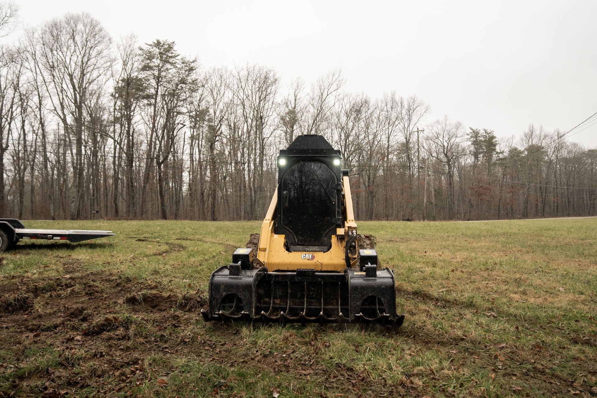 Yellow skid steer with a shredder attachment on a field, forest in background.