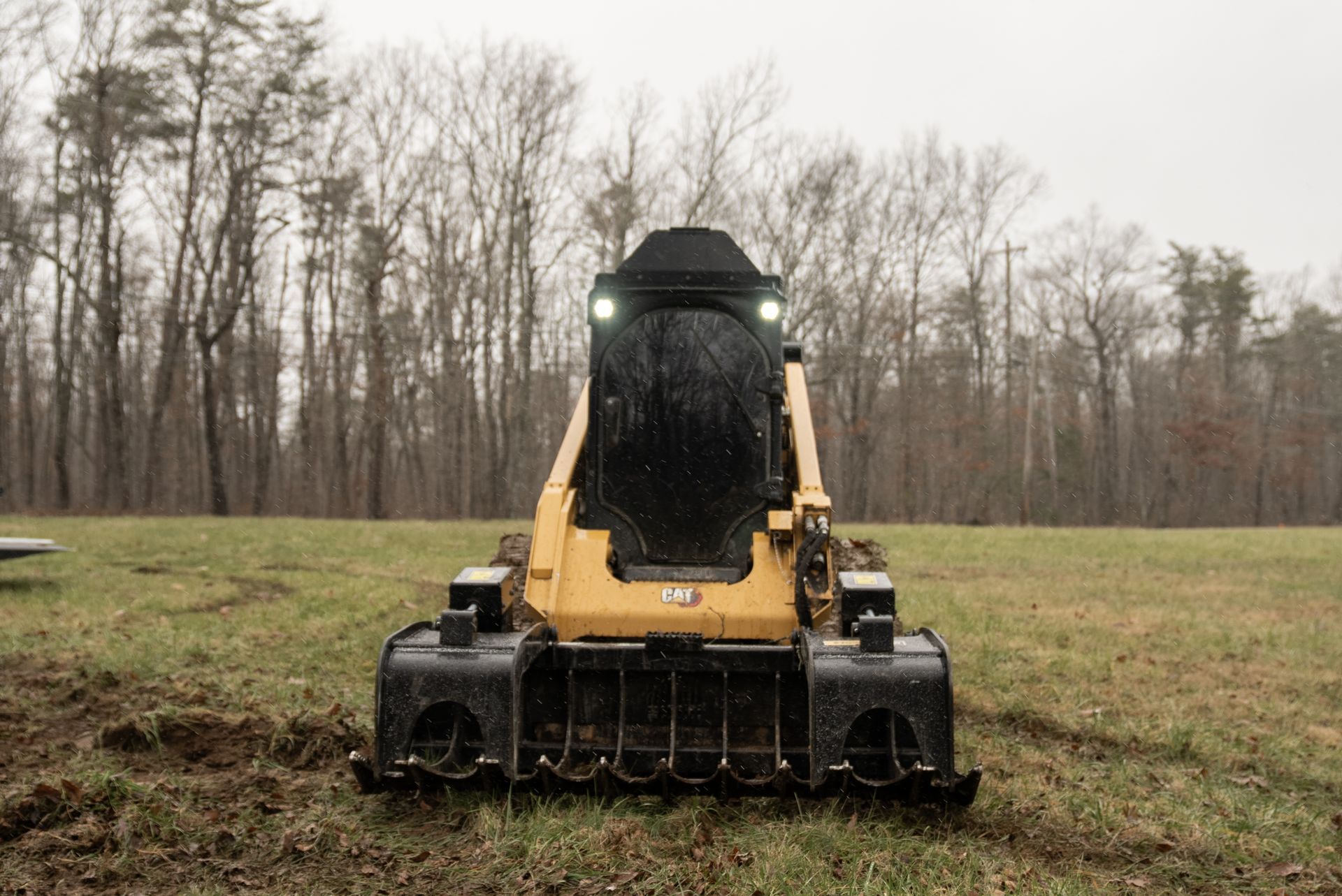 Yellow skid steer with front attachment on muddy field, trees in the background, possibly rainy day.