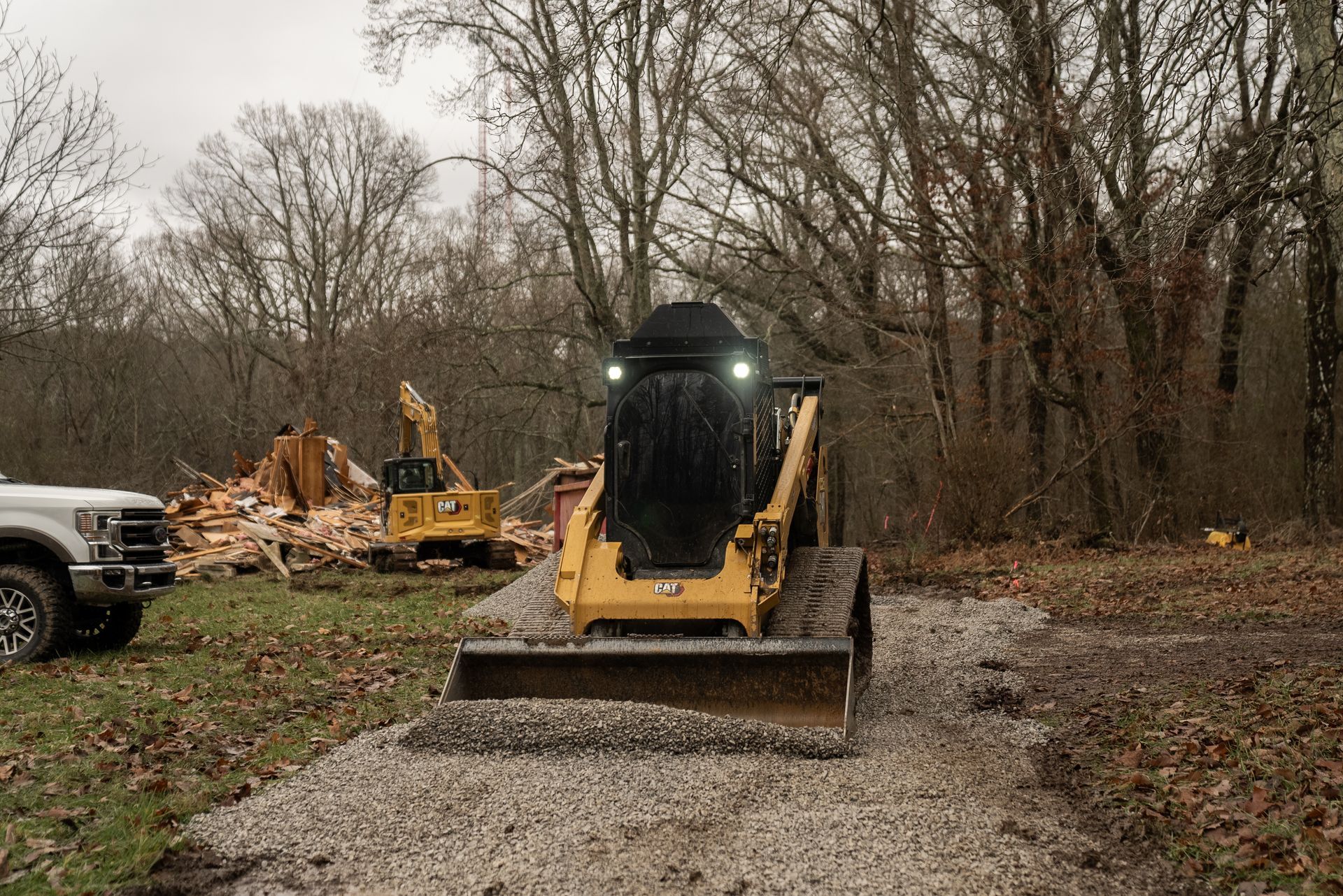 Skid steer loader on gravel path, another excavator demolishing a building in background. Cloudy day.