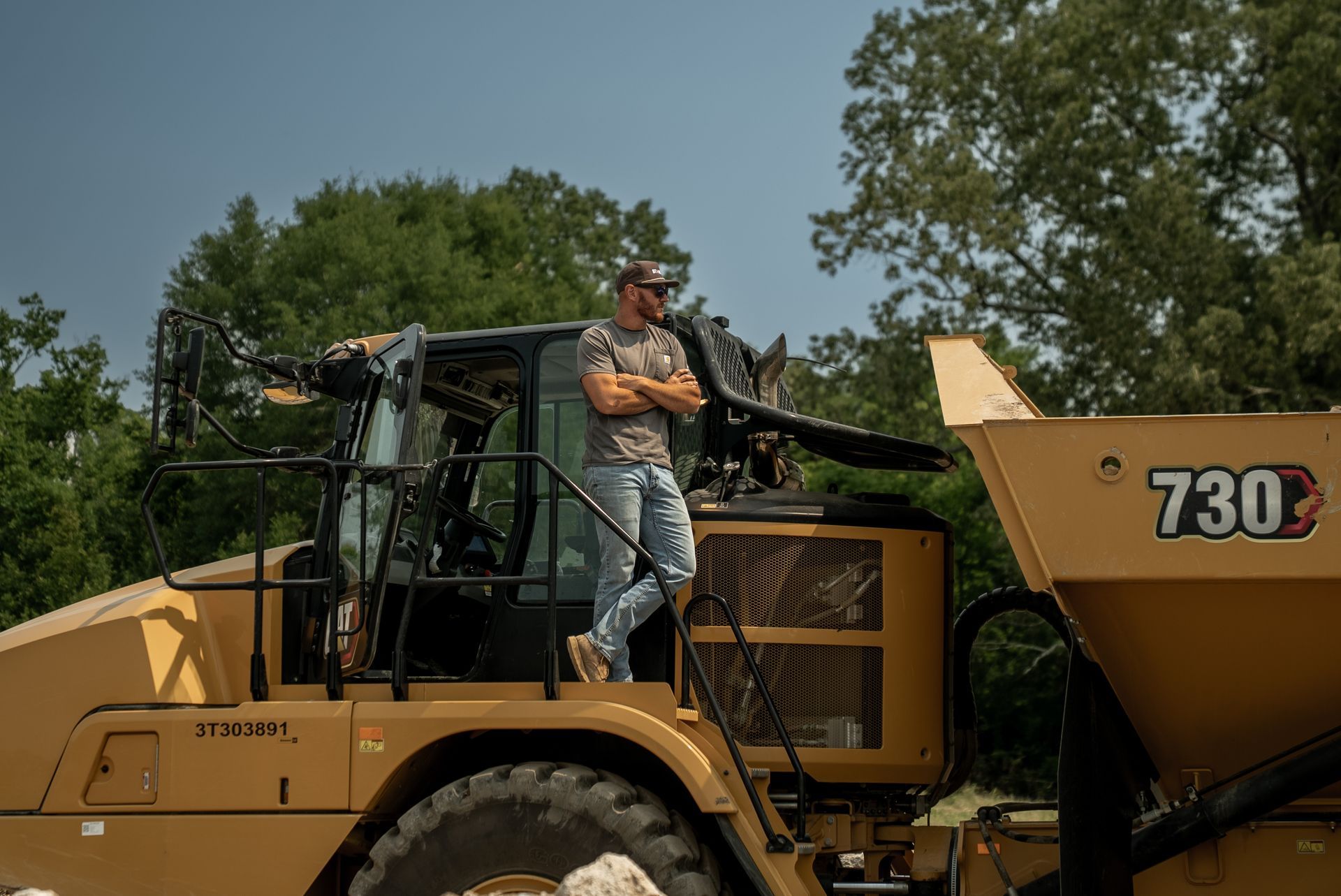 Man standing on a yellow 730B Caterpillar hauler in a wooded area, arms crossed.