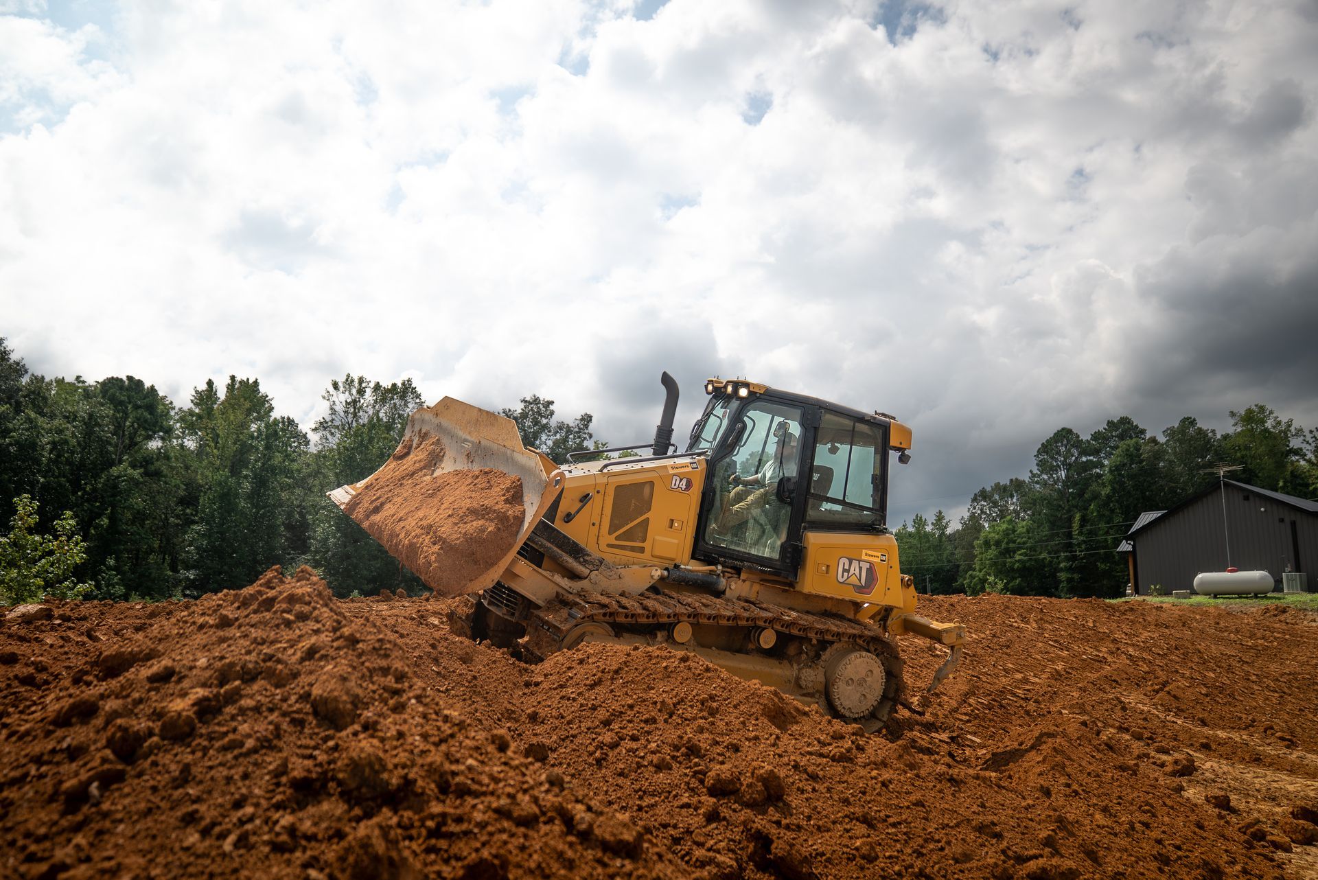Yellow bulldozer pushing dirt on a construction site; trees and a cloudy sky in the background.