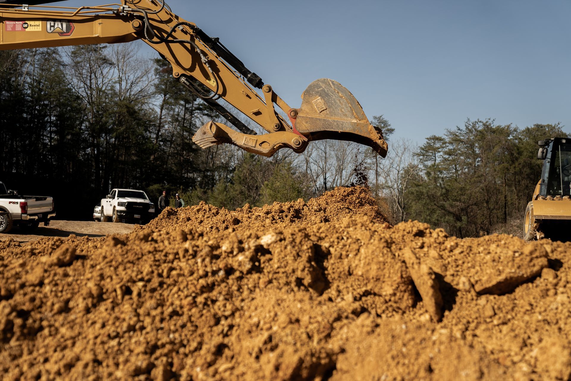 Excavator arm lifting dirt from a pile at a construction site with vehicles and trees in the background.