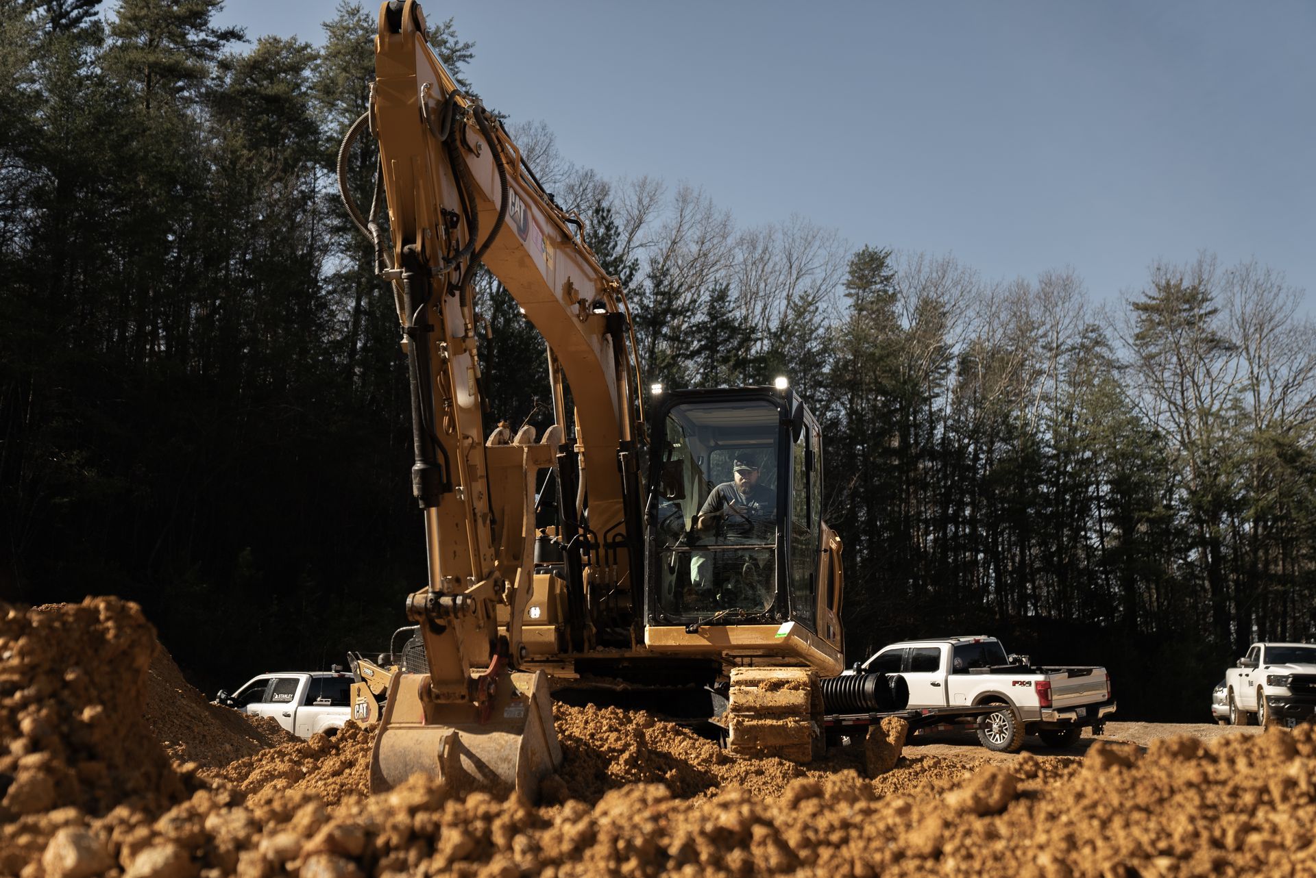 Yellow excavator digging into dirt; white trucks parked nearby; trees in background.