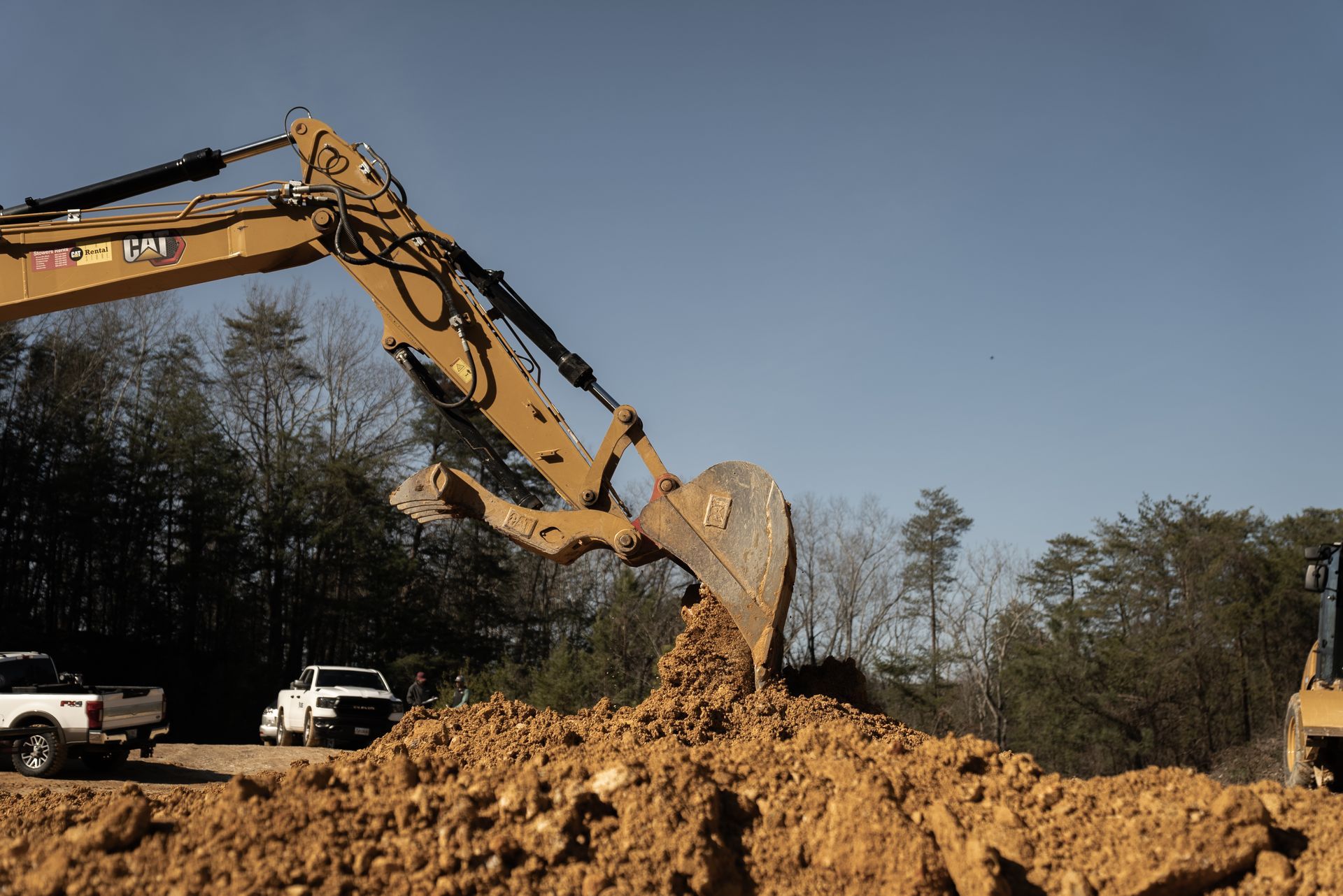 Excavator digging dirt at a construction site with white trucks and trees in the background under a blue sky.