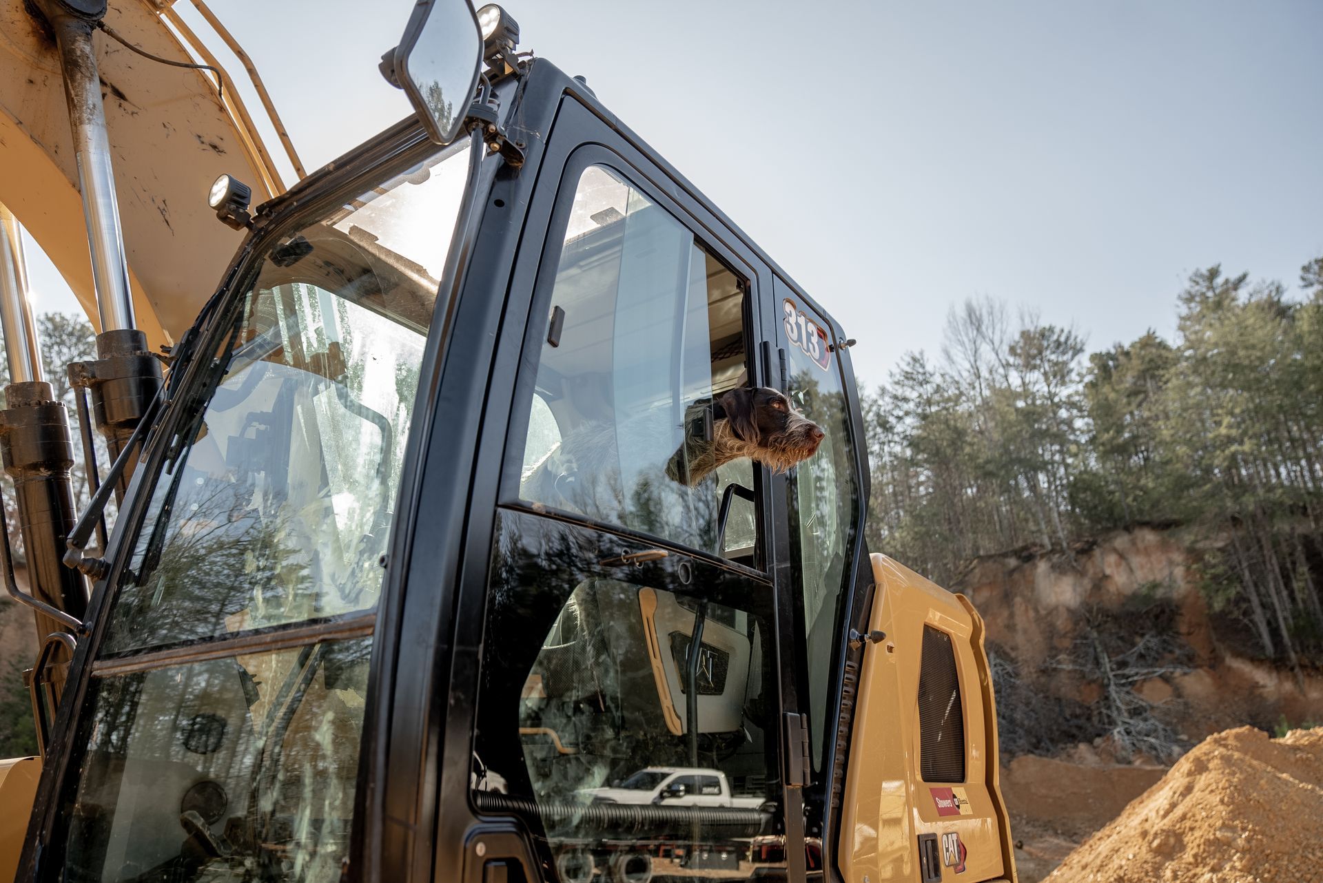 Excavator cab with operator visible; outdoors, bright sunlight.