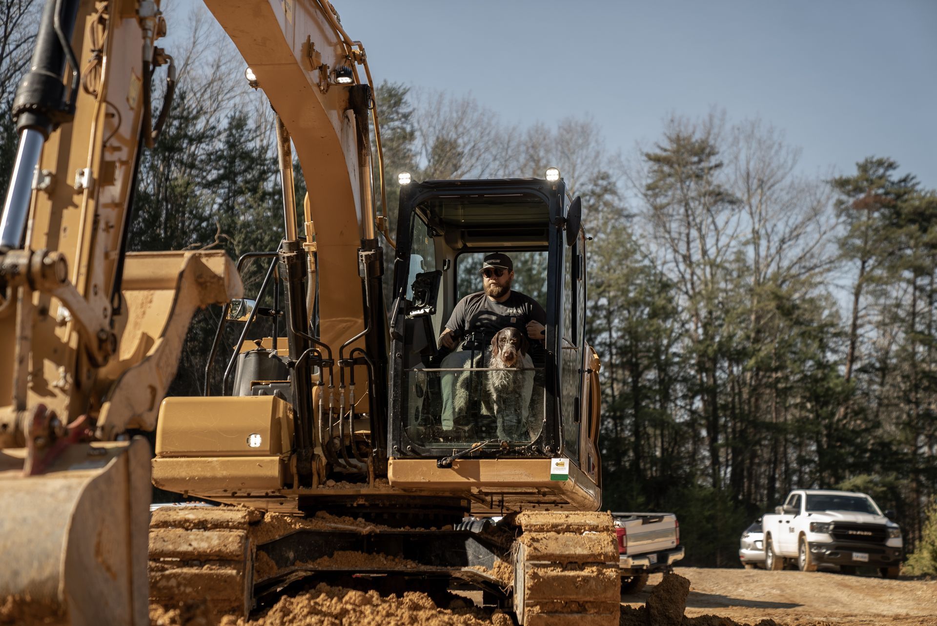 A person operates an excavator at a construction site near trees and a parked truck on a sunny day.