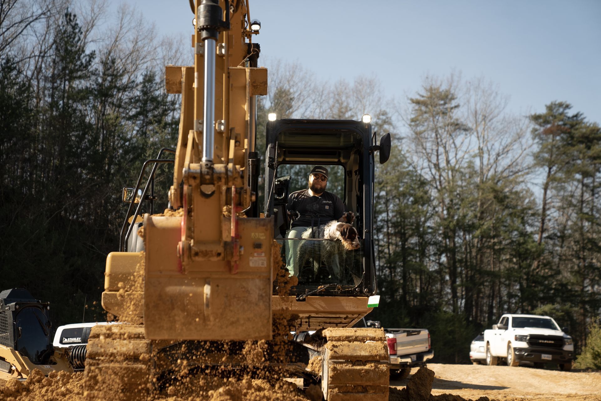 Excavator operator in cabin, digging dirt; white trucks parked nearby on sunny construction site.