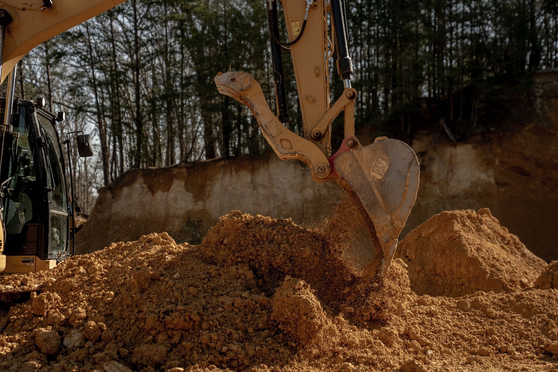 An excavator scoops up brown soil at a construction site near a forest.