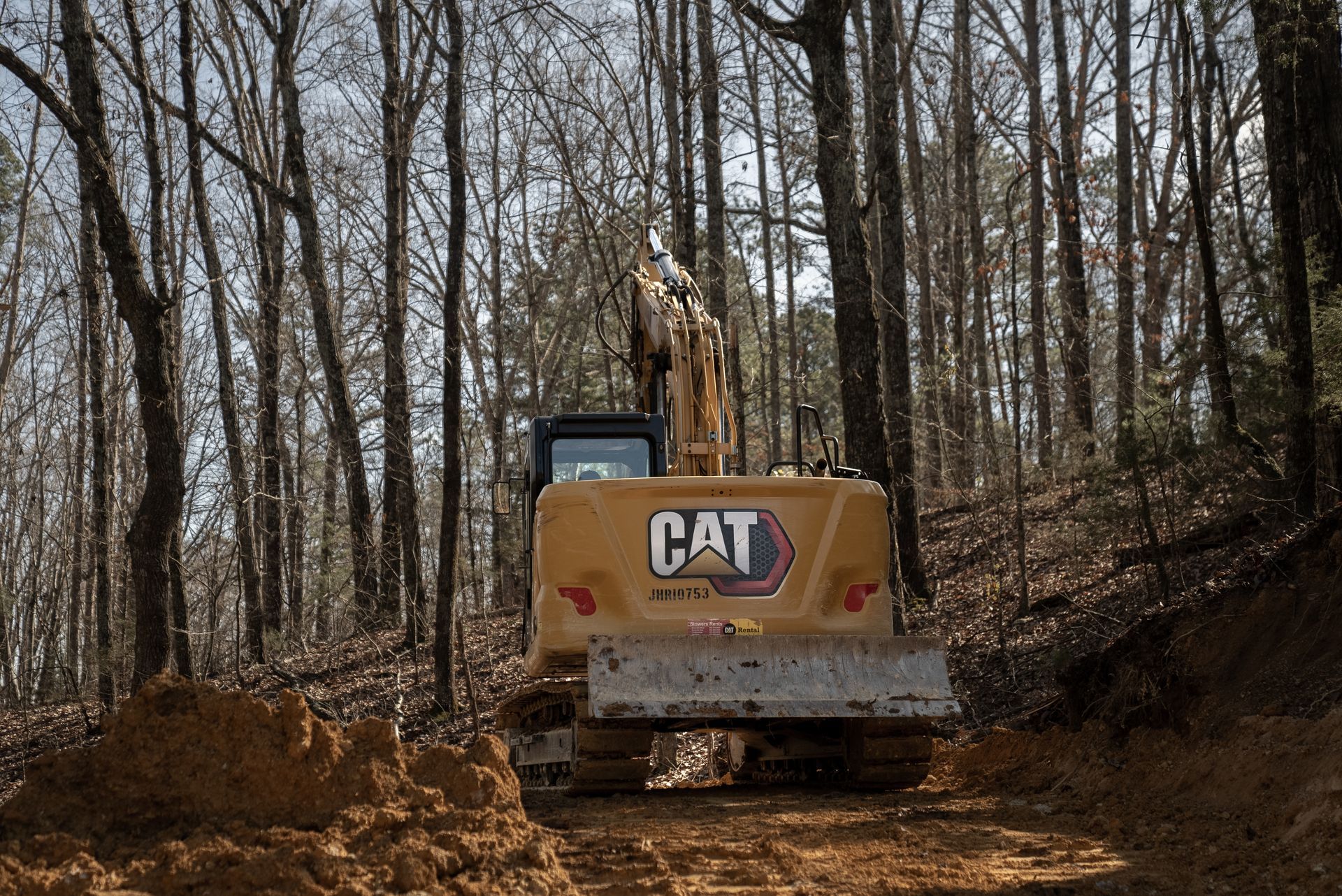 Yellow CAT excavator clears a muddy path in a wooded area, trees in the background.