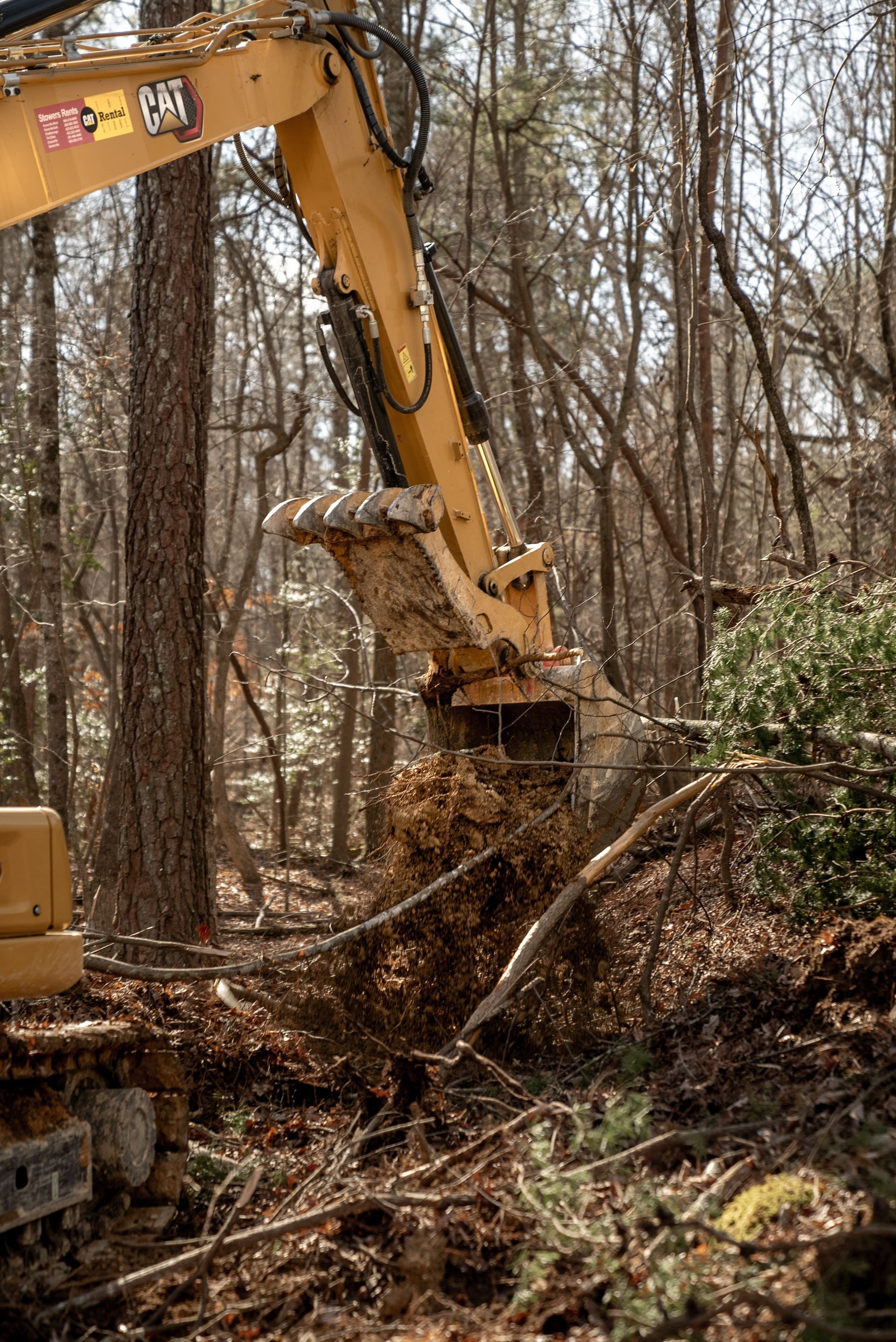 Yellow excavator bucket digging into soil in a wooded area, clearing land.