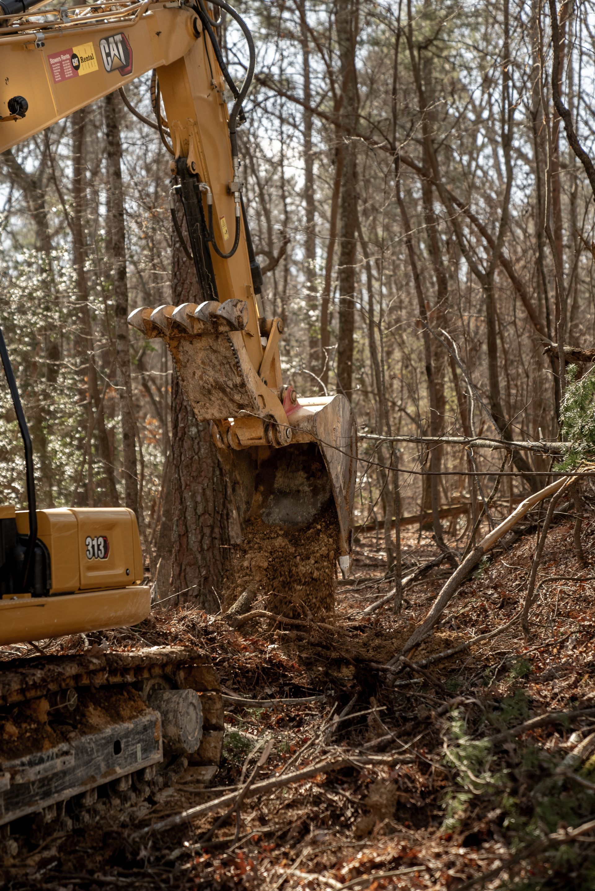 Yellow excavator bucket moving dirt and debris in a wooded area.