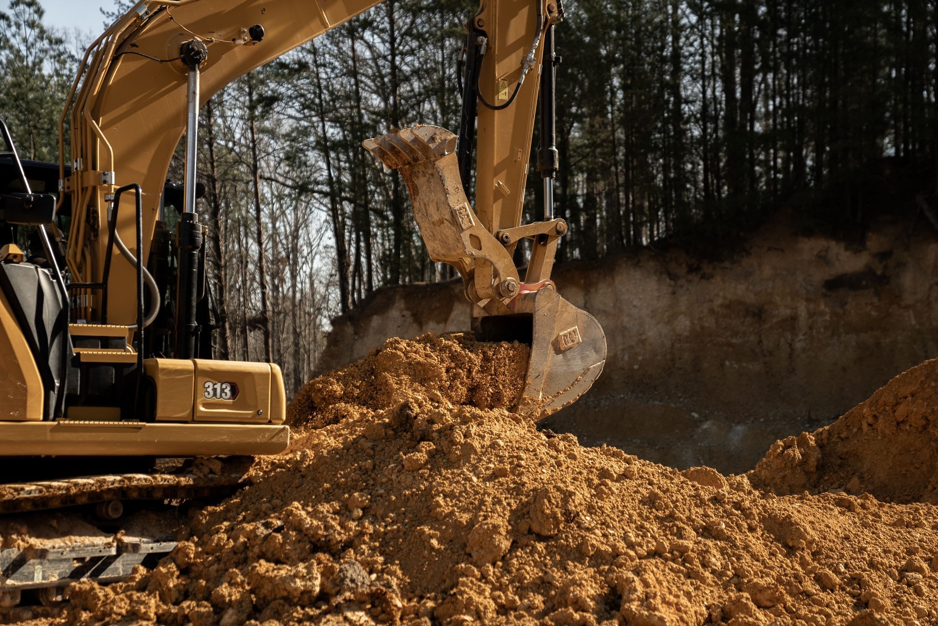 Yellow excavator digging brown soil outdoors near trees.