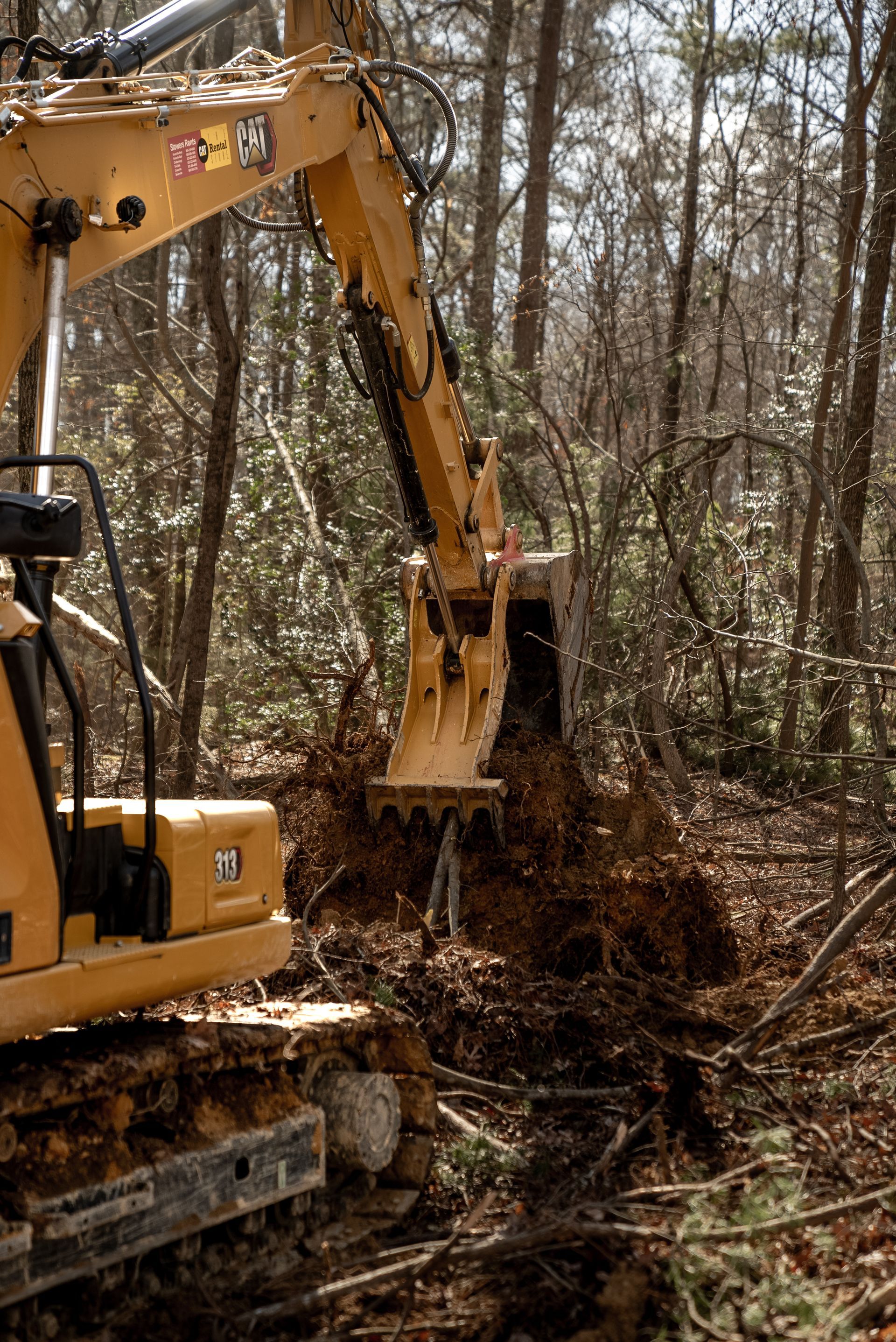 Yellow excavator digging in a wooded area, scooping up dirt and roots.