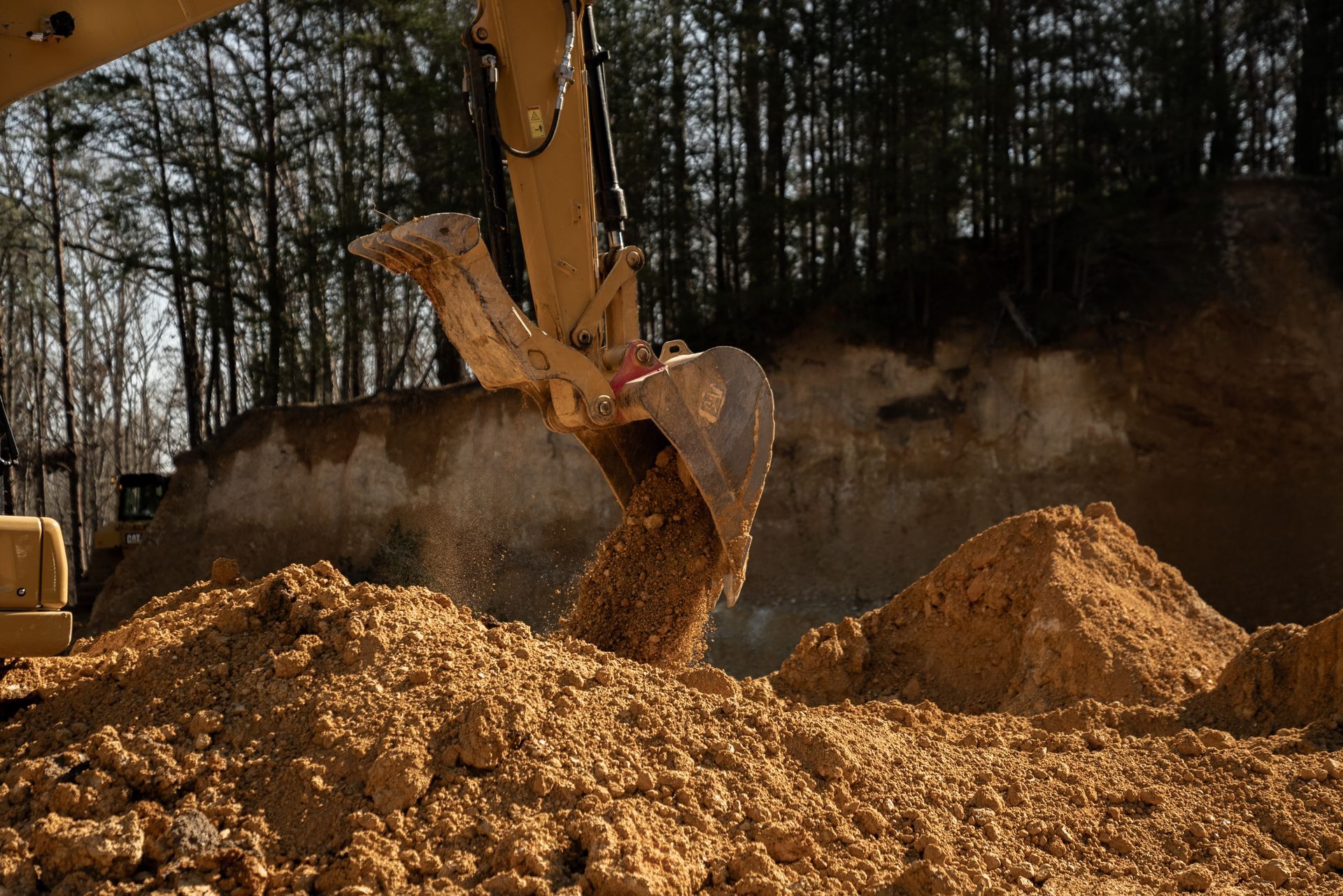 Excavator scooping dirt at a construction site, brown dirt piles, trees in background.