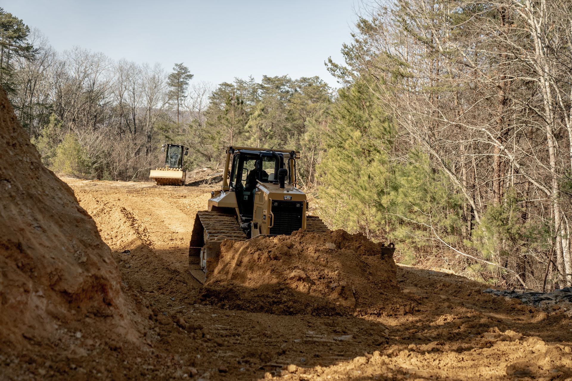 Bulldozer pushing dirt on a muddy road, with another construction vehicle in the distance, trees line the sides.