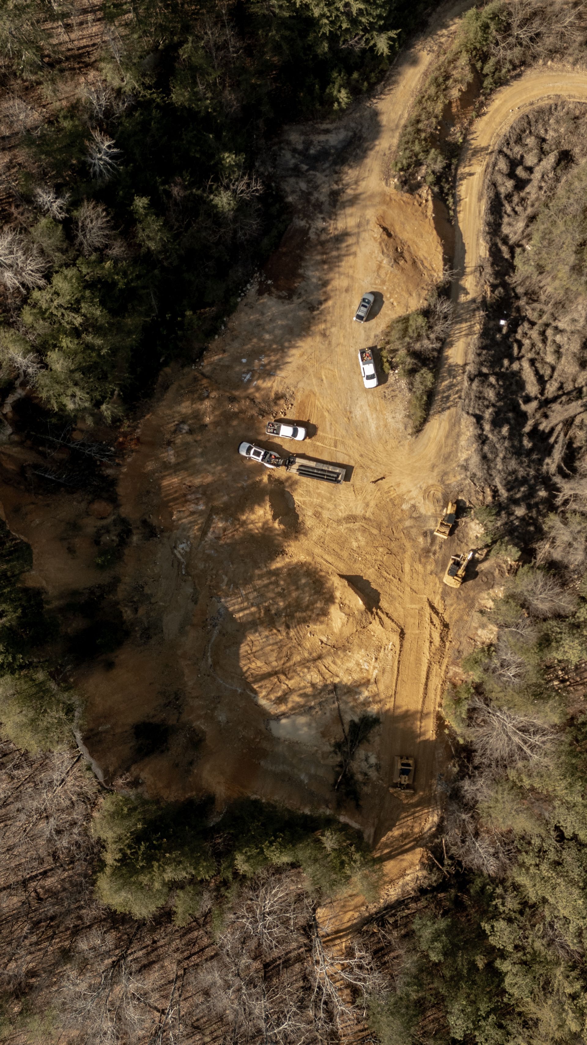 Aerial view of a dirt road and clearing in a forest, with several vehicles and earthmoving equipment.