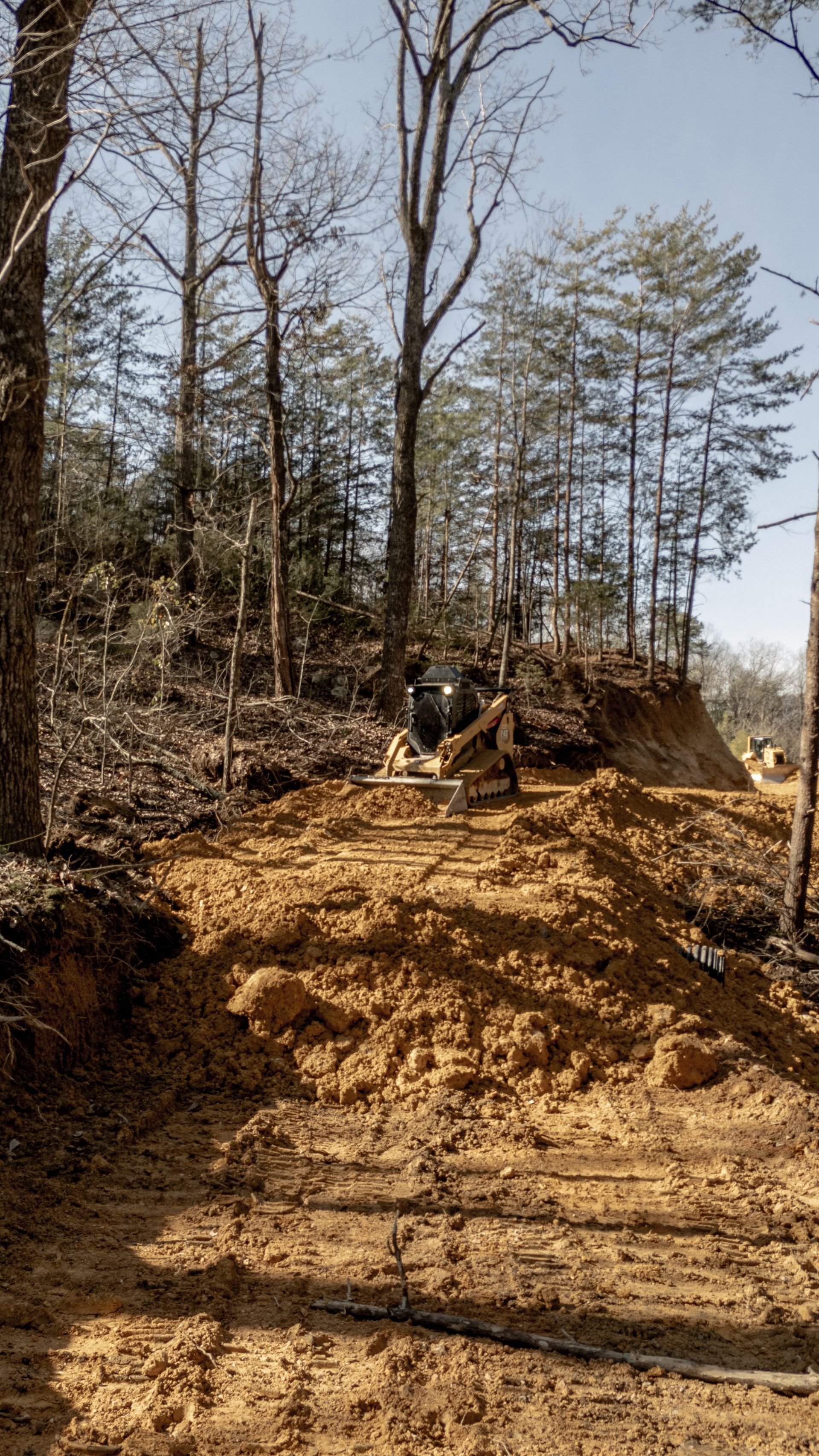Dirt road through a construction site, with trees in the background. A person stands near a pile of dirt.