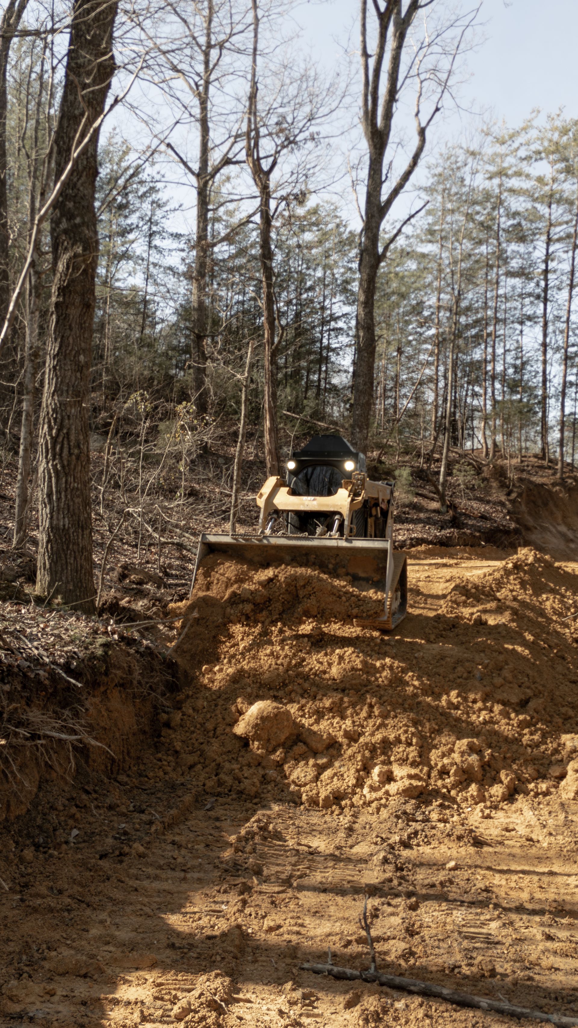 A yellow tractor with a blade levels a hillside of brown dirt in a wooded area.