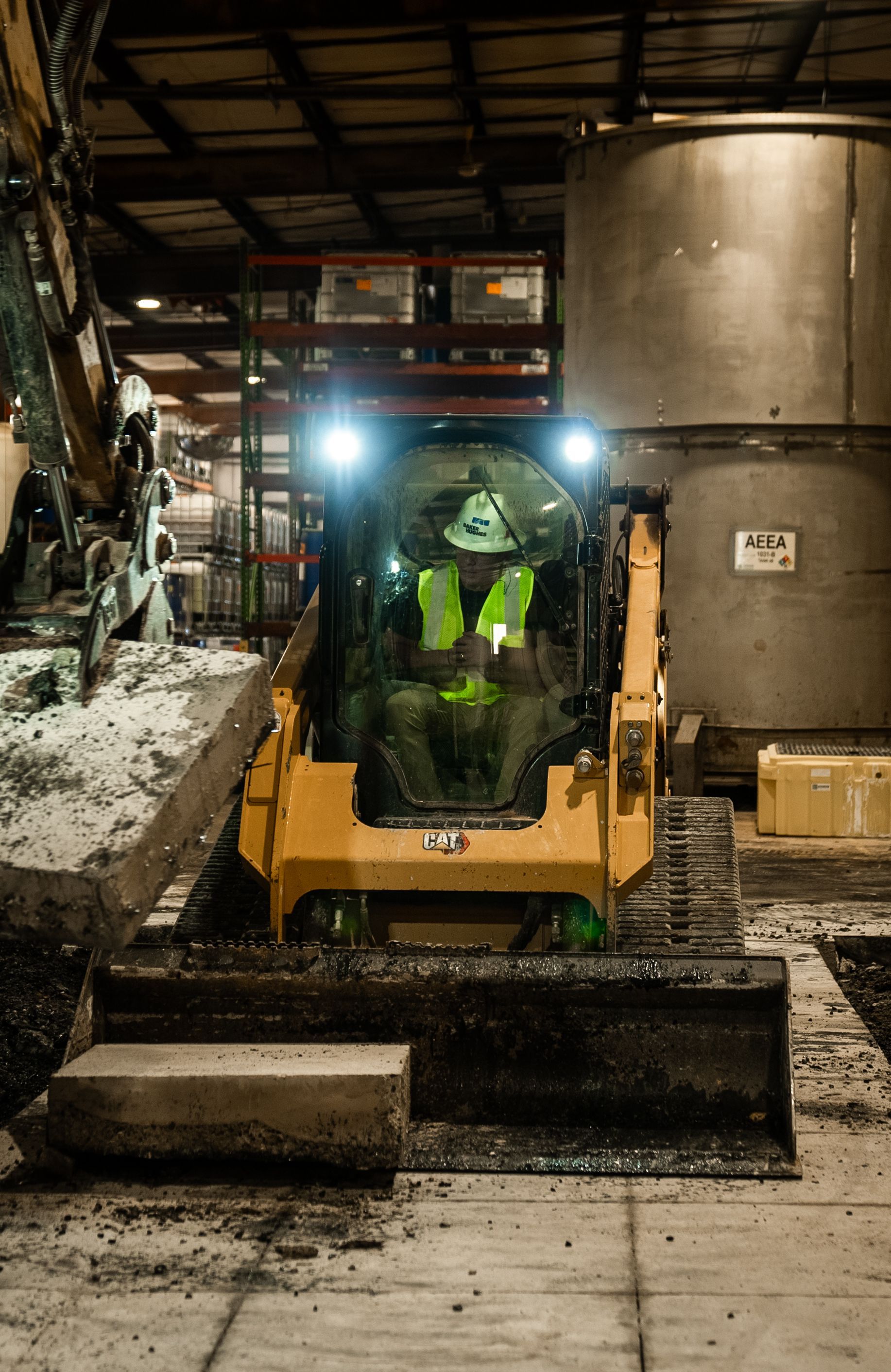 Yellow Caterpillar skid steer operating indoors, moving debris. Driver wearing a reflective vest.