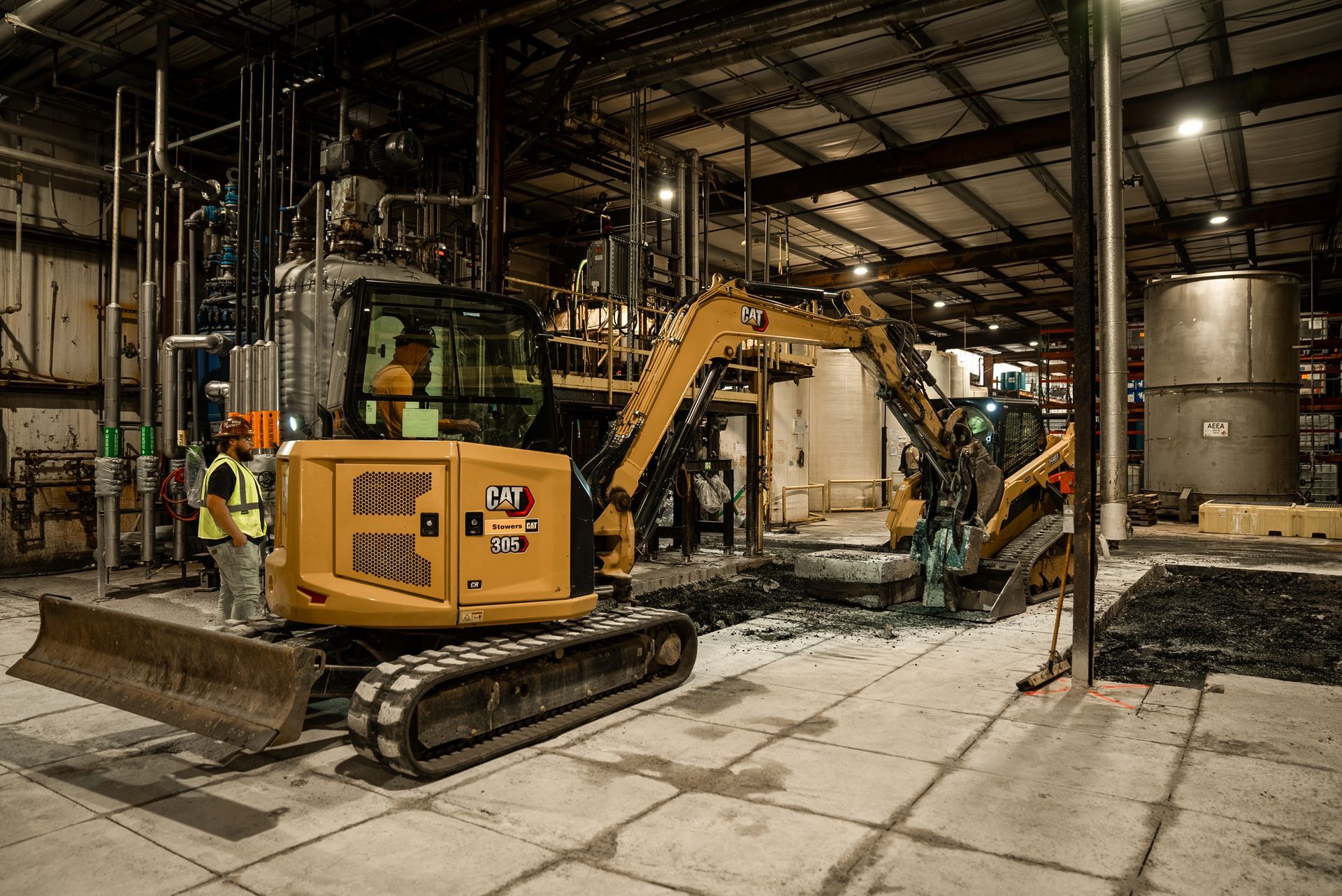 Yellow excavator inside a factory, worker observing, pipes and tanks in the background.