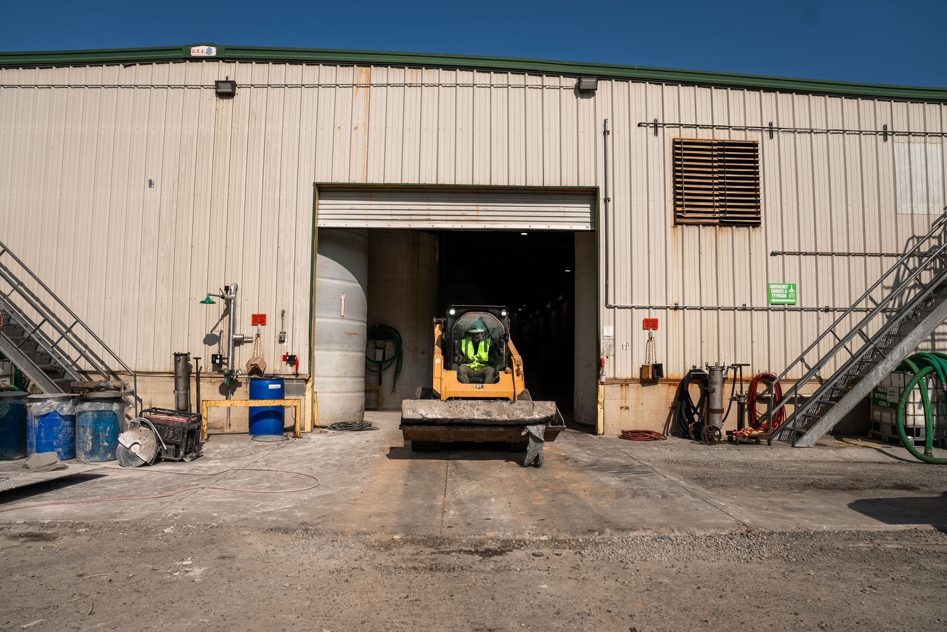 Yellow skid steer exiting a warehouse door on a gravel surface. Barrels and equipment visible nearby.