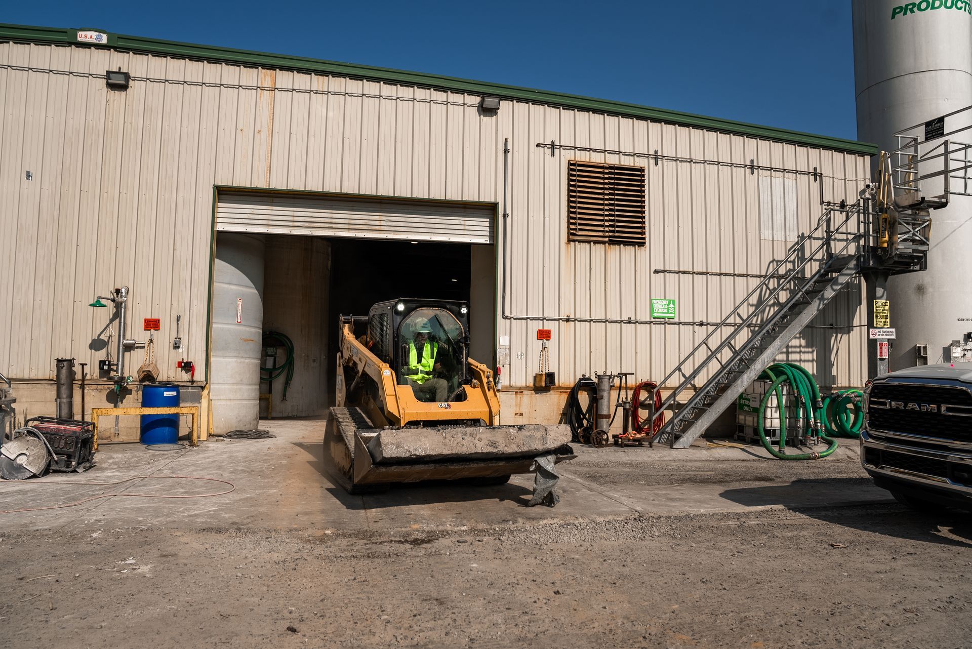 A skid steer loader exits a warehouse. Exterior shot with blue sky, concrete, and industrial equipment.