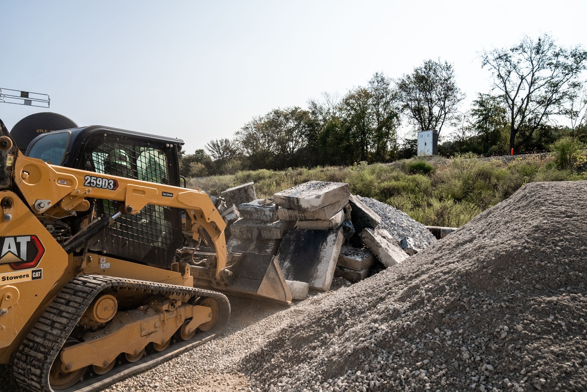 Yellow CAT skid steer pushing concrete debris into a gravel pile, with trees in the background.