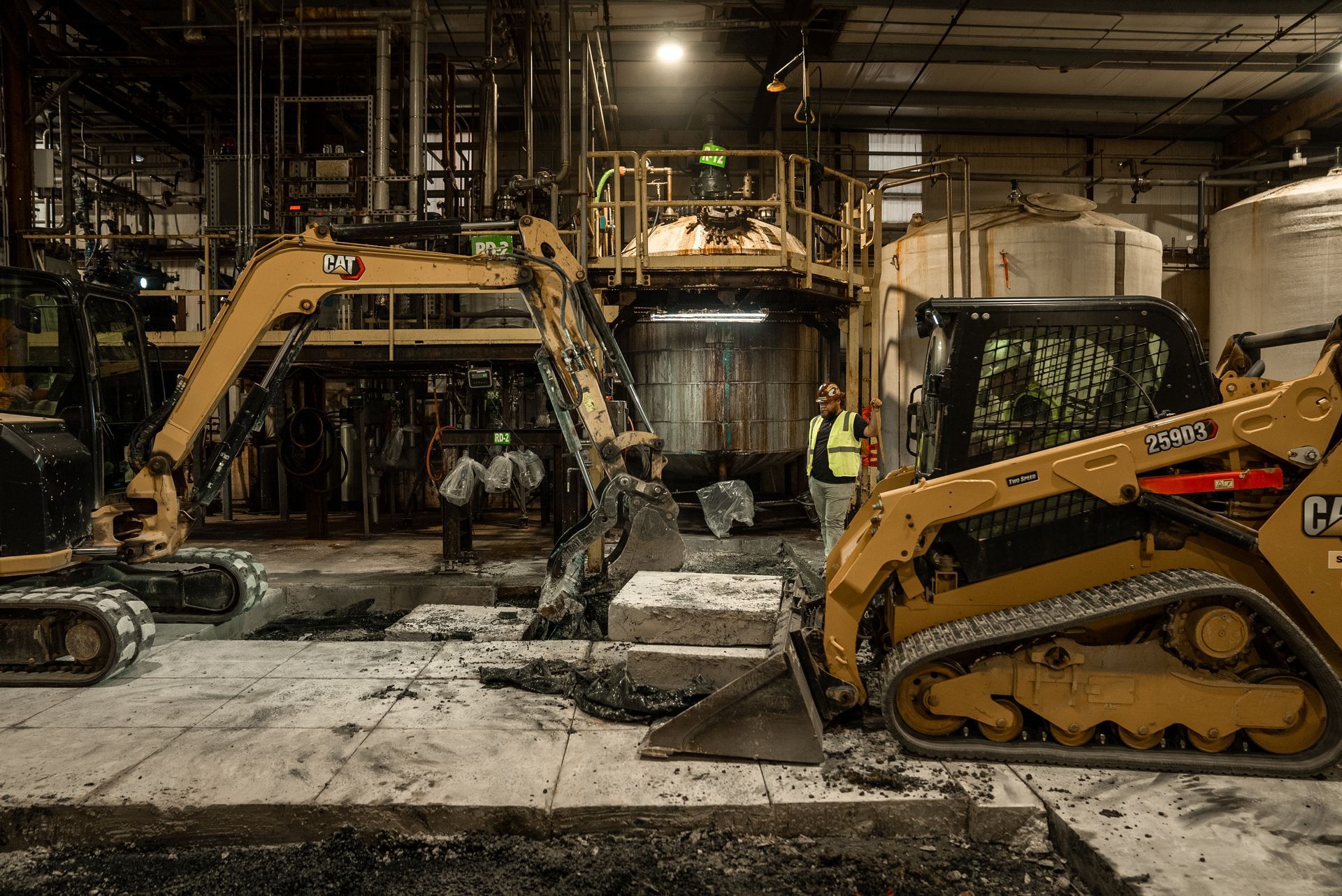 Heavy machinery working inside an industrial building. Yellow excavators and a skid steer operate on a concrete floor.