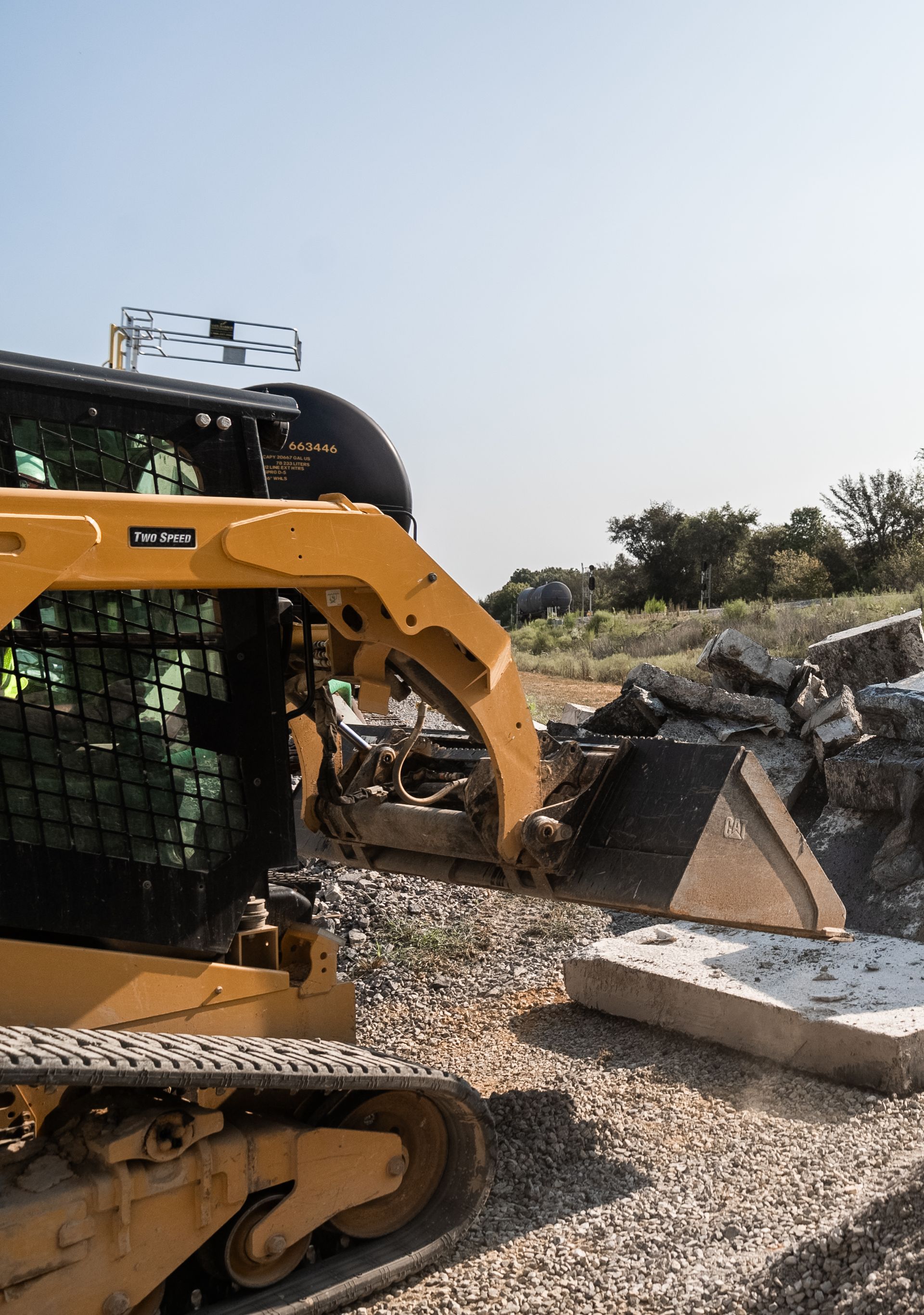 Yellow skid steer with grapple tearing up concrete in a gravel lot under a blue sky.