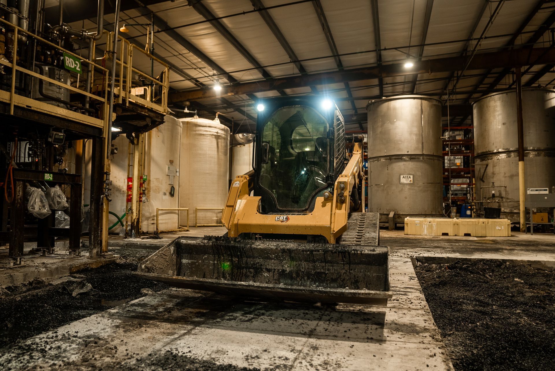 Yellow skid steer inside industrial building, moving dark material. Large tanks and metal structure visible.