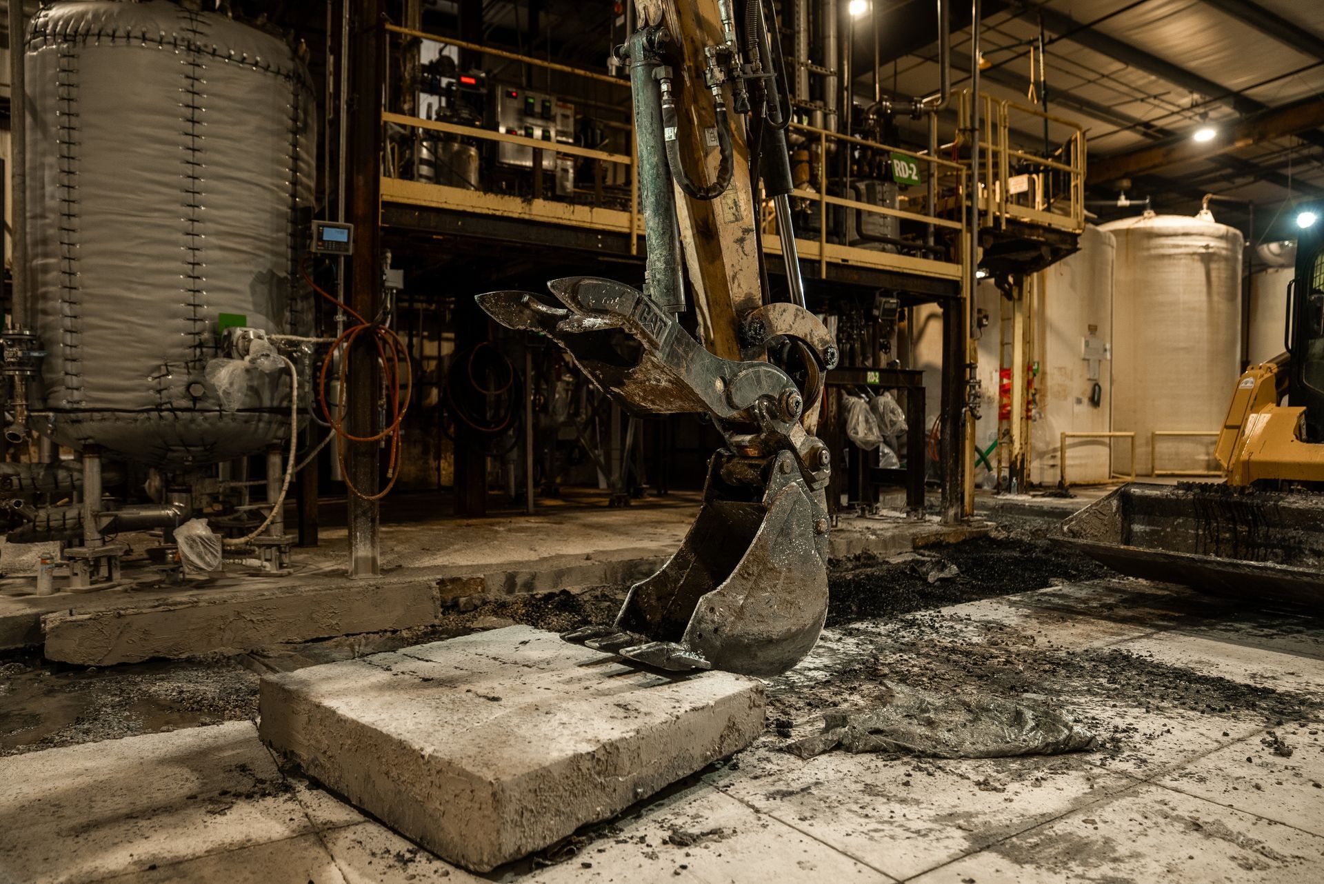 Excavator in industrial setting breaking concrete slab; tanks and metal structures in background.