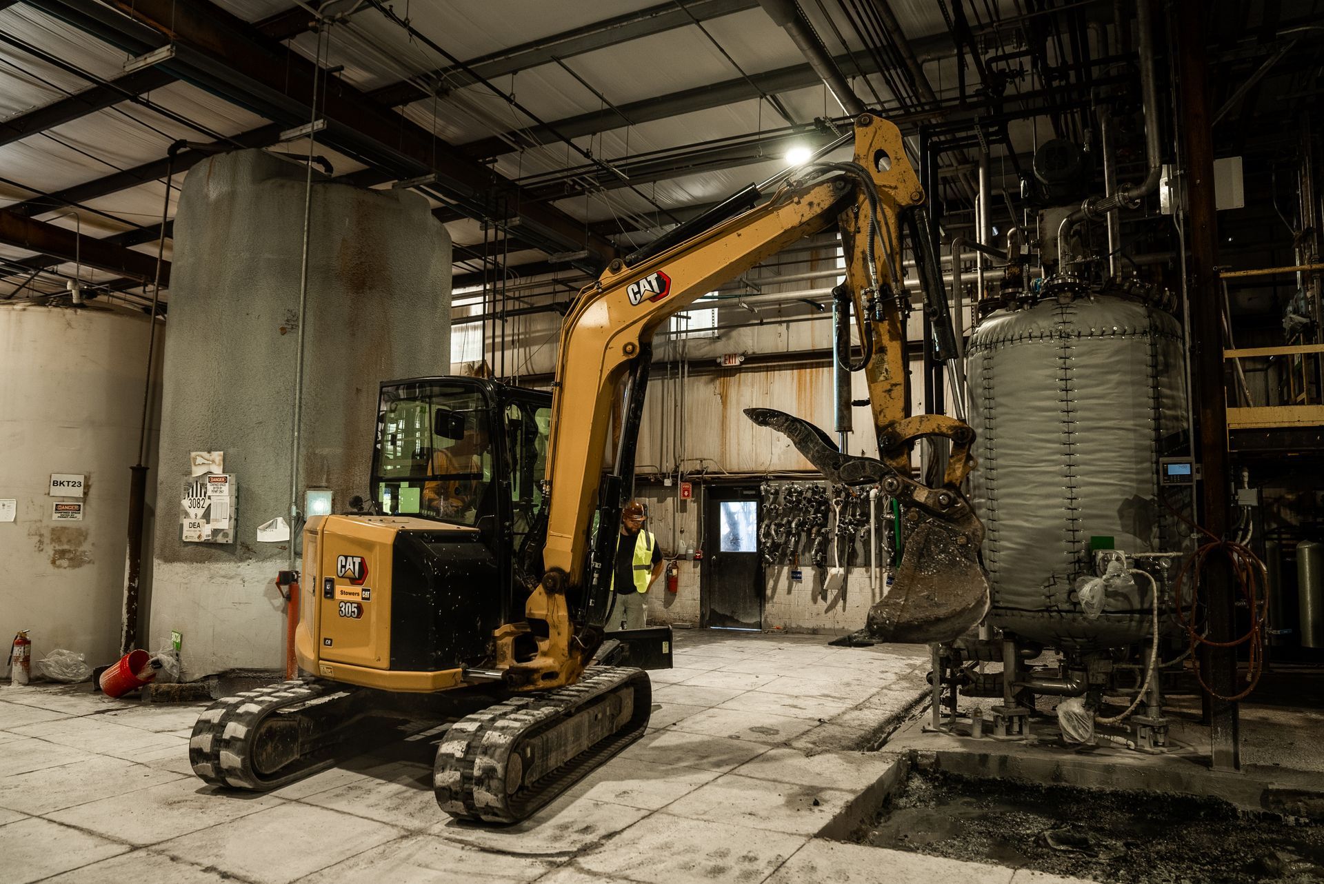 Yellow excavator inside a factory, working near cylindrical tanks. A worker stands nearby.