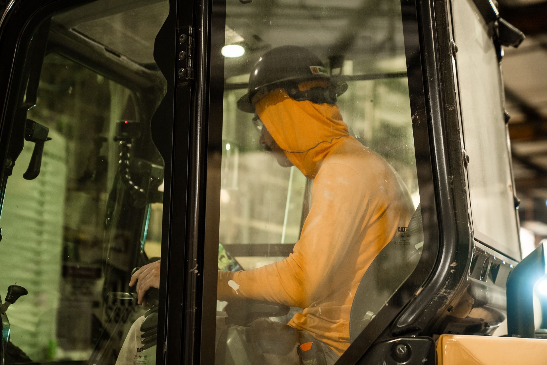 Person operates machinery inside a cab, wearing a hat and orange protective gear.