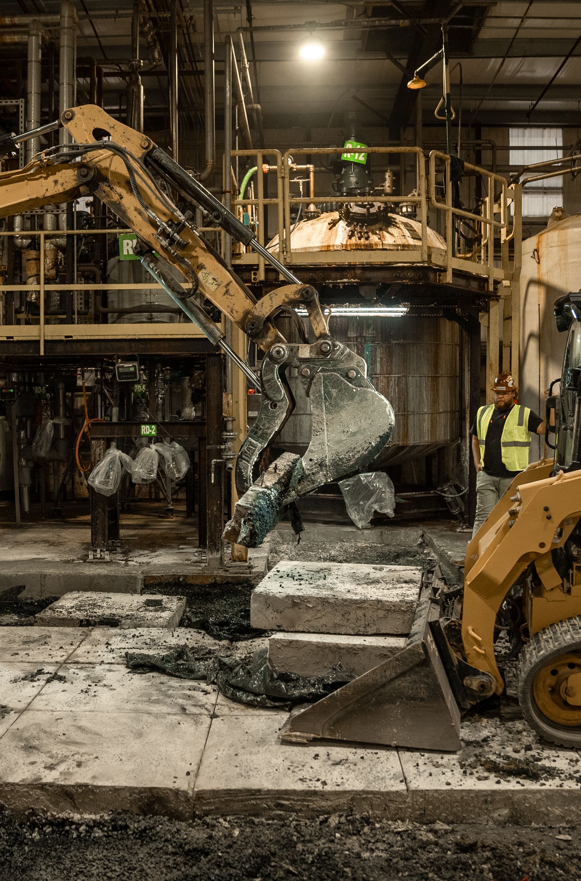 Excavator scoops up metal debris in a factory, with workers nearby.