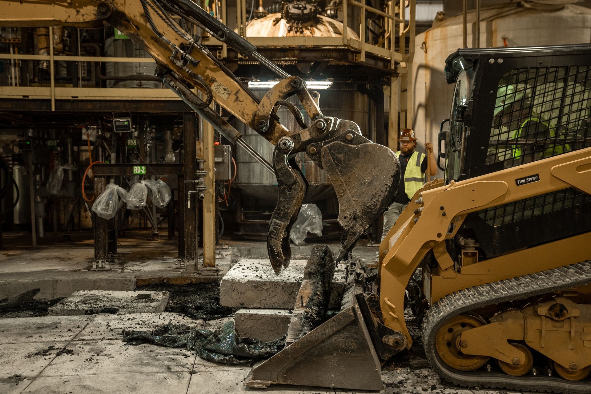 Yellow skid steer with claw demolishes concrete blocks in an industrial setting, worker in vest watches.