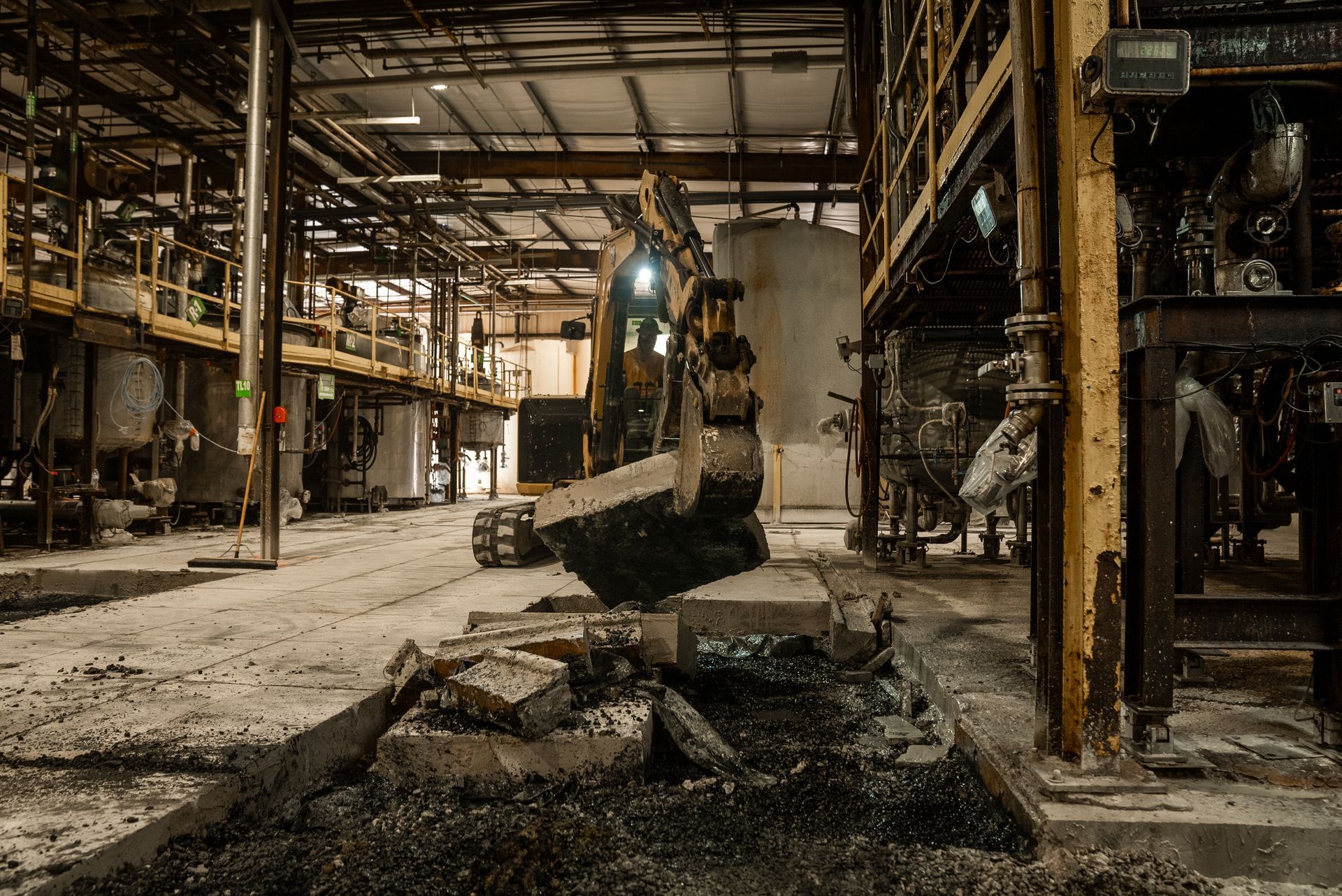 Excavator in an industrial facility, moving debris. The interior is dark and shows signs of damage.