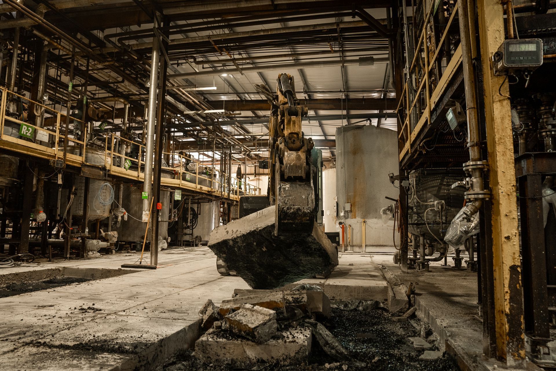 An excavator demolishes a factory interior. Debris covers the floor, metallic structures and tanks visible.