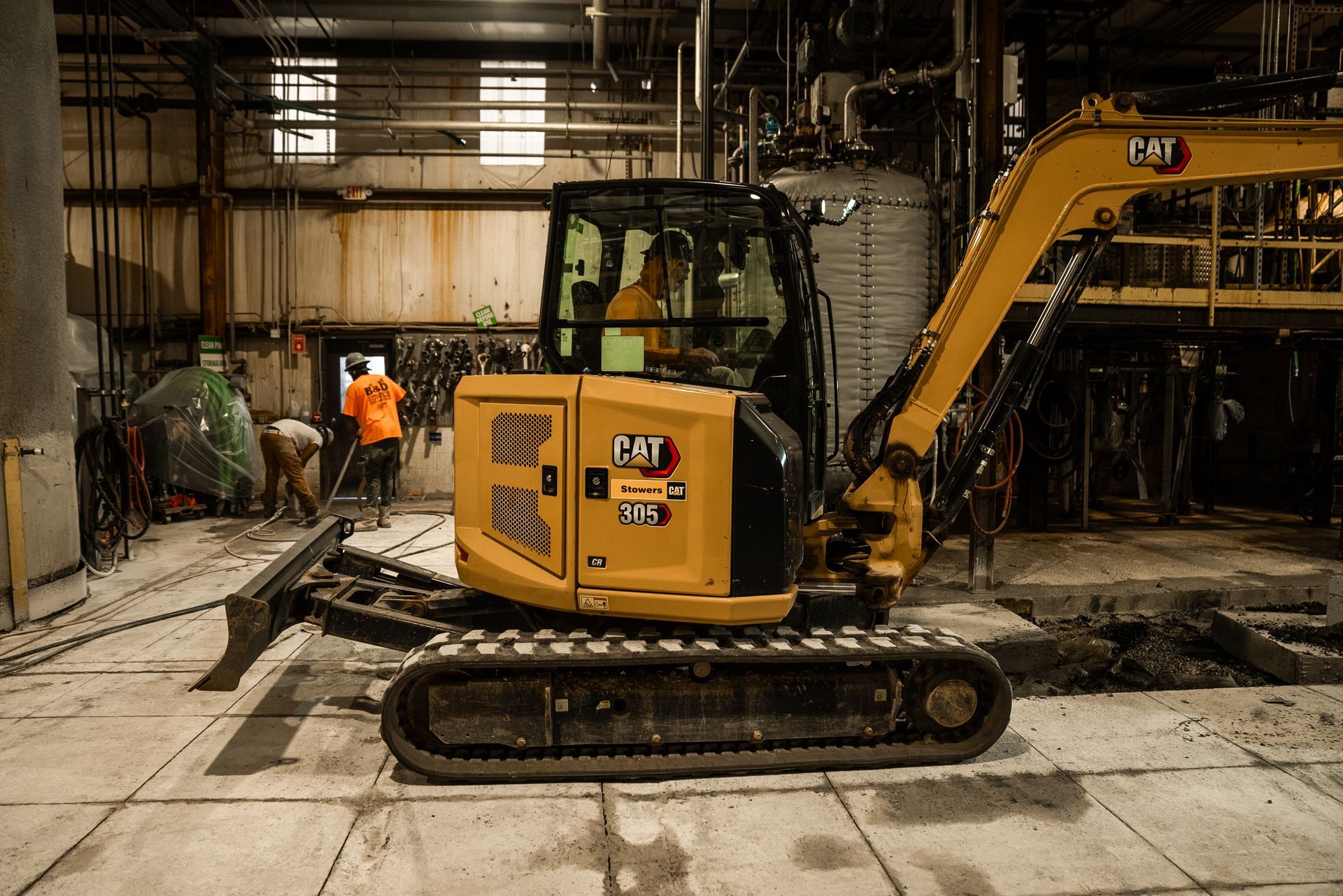 Yellow Caterpillar excavator inside a building; workers in background.